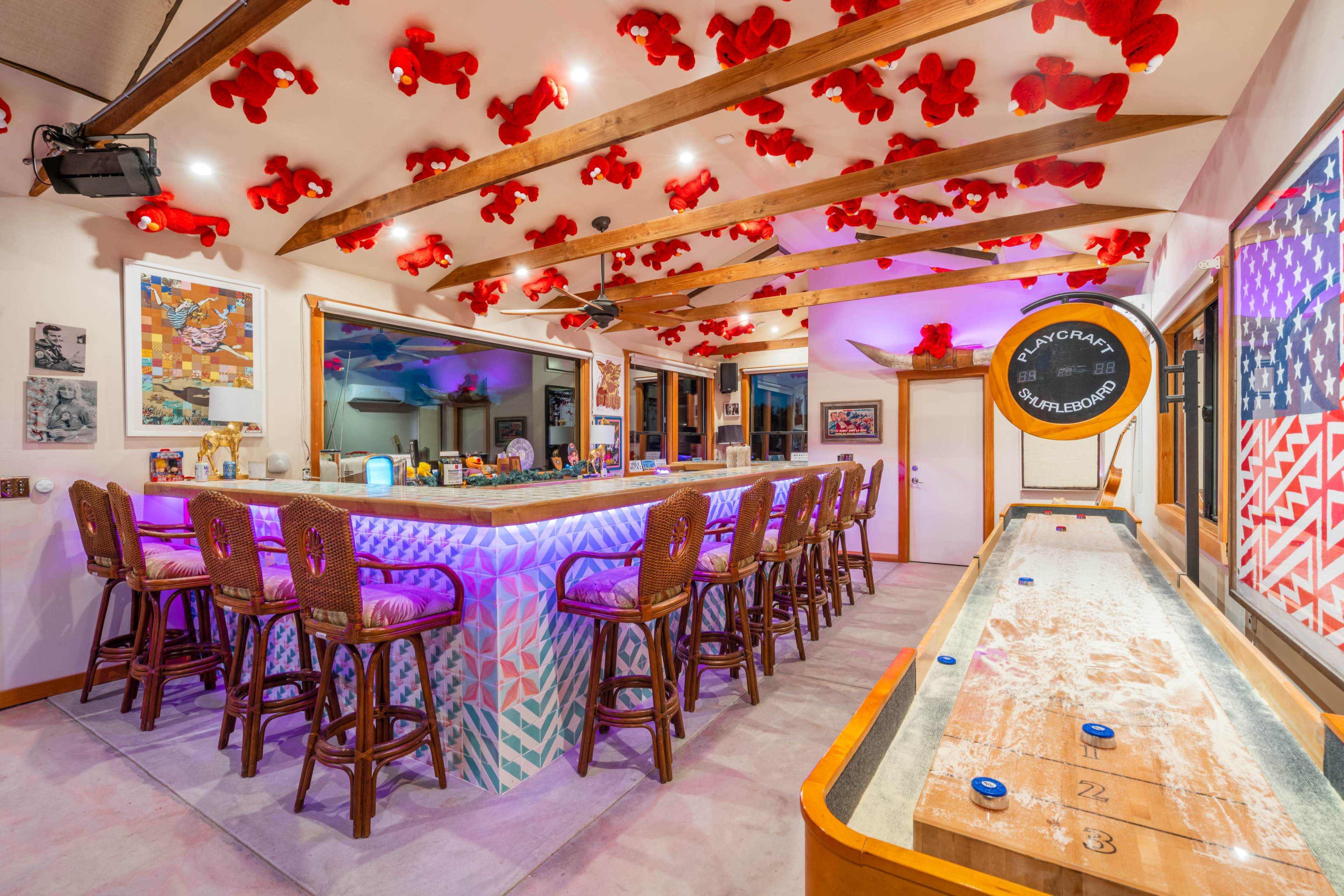 A brightly lit bar area with a long countertop, high chairs, and a shuffleboard table, all decorated with red teddy bears hanging from the ceiling.