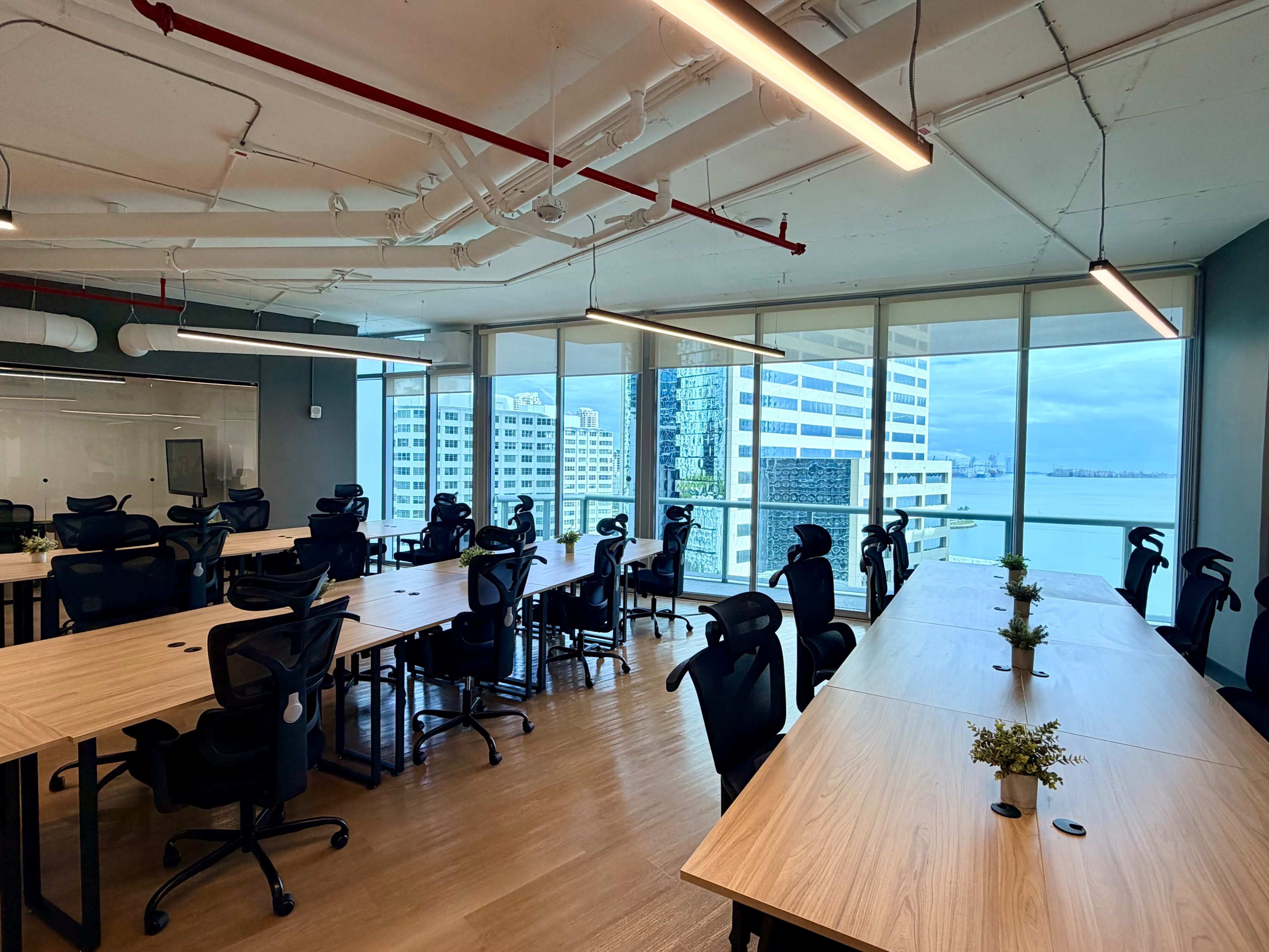 The image shows a modern office space with multiple wooden desks and ergonomic chairs, large windows, and a view of a cityscape and waterfront.