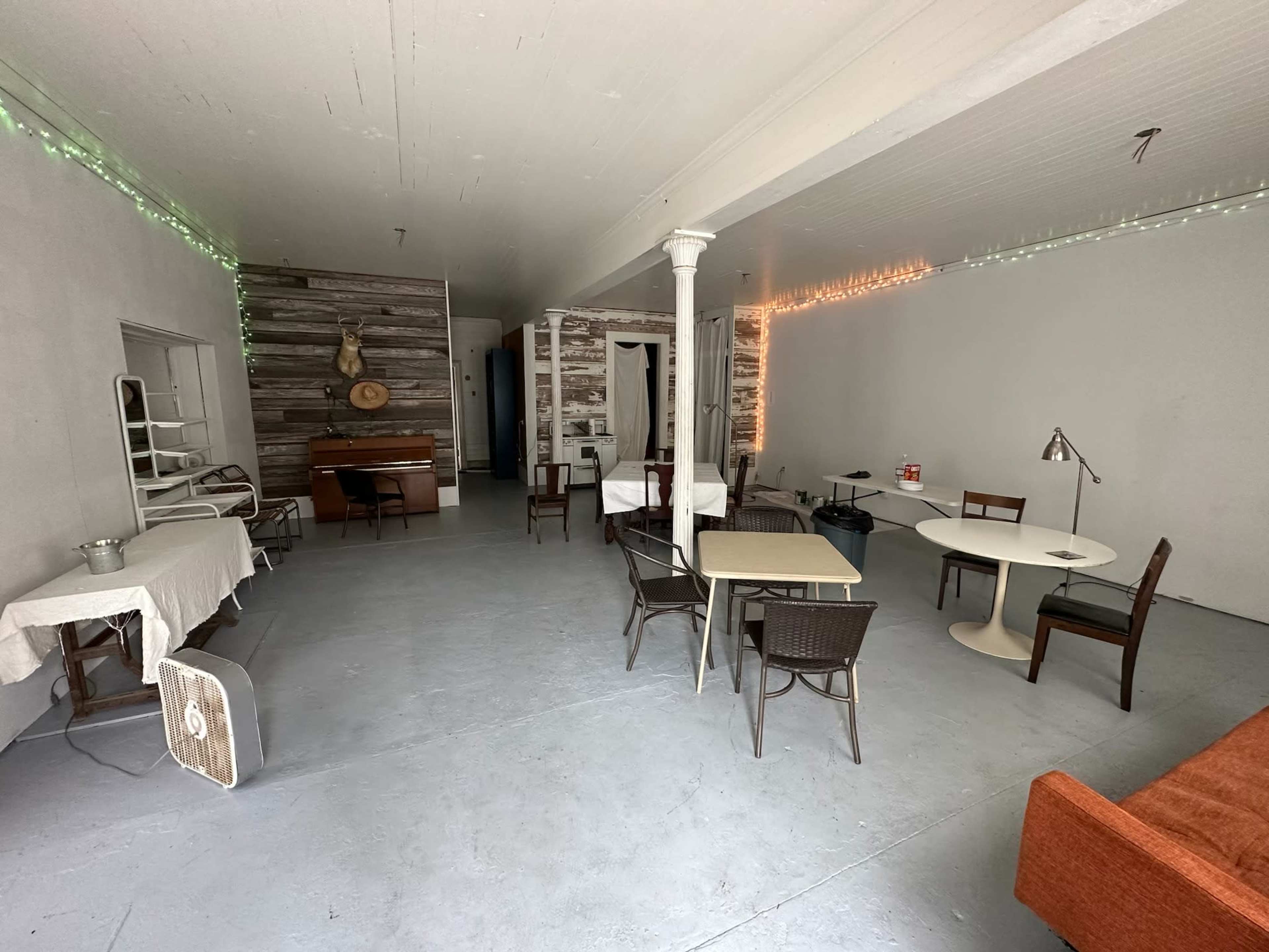 The image shows a spacious, empty interior with various tables and chairs, a piano in the background, and string lights along the walls.