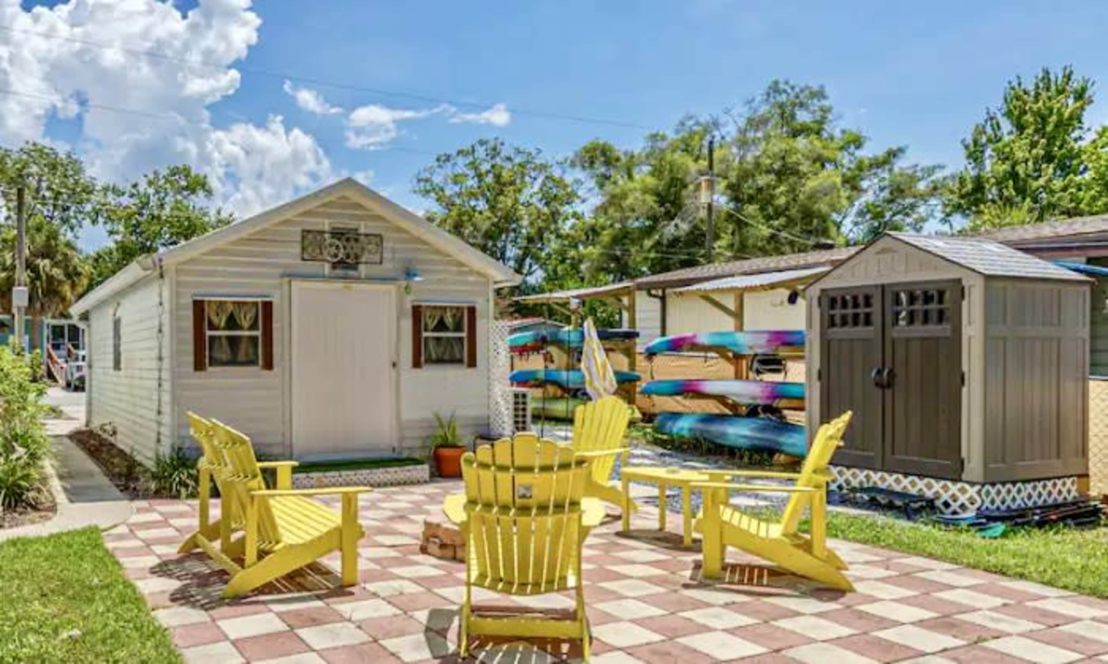 A backyard with yellow Adirondack chairs arranged around a square brick patio, flanked by a small white shed and a storage unit, while colorful kayaks are neatly stacked nearby.