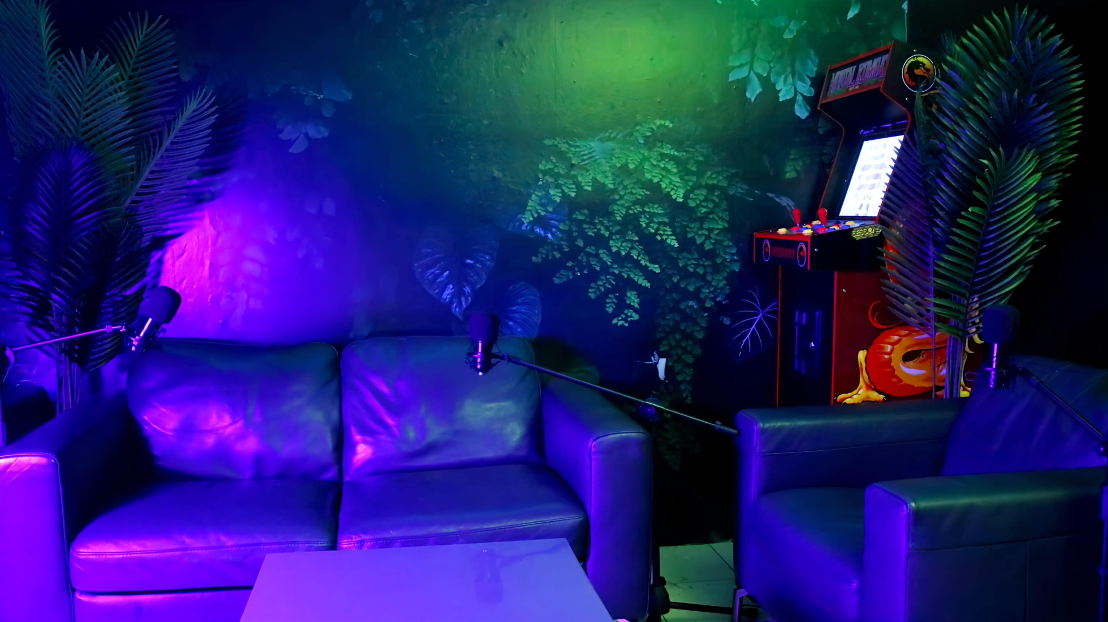 The scene features a dimly lit room with a dark green plant-themed mural, a blue leather couch, a white table, and a vintage arcade machine in the corner.