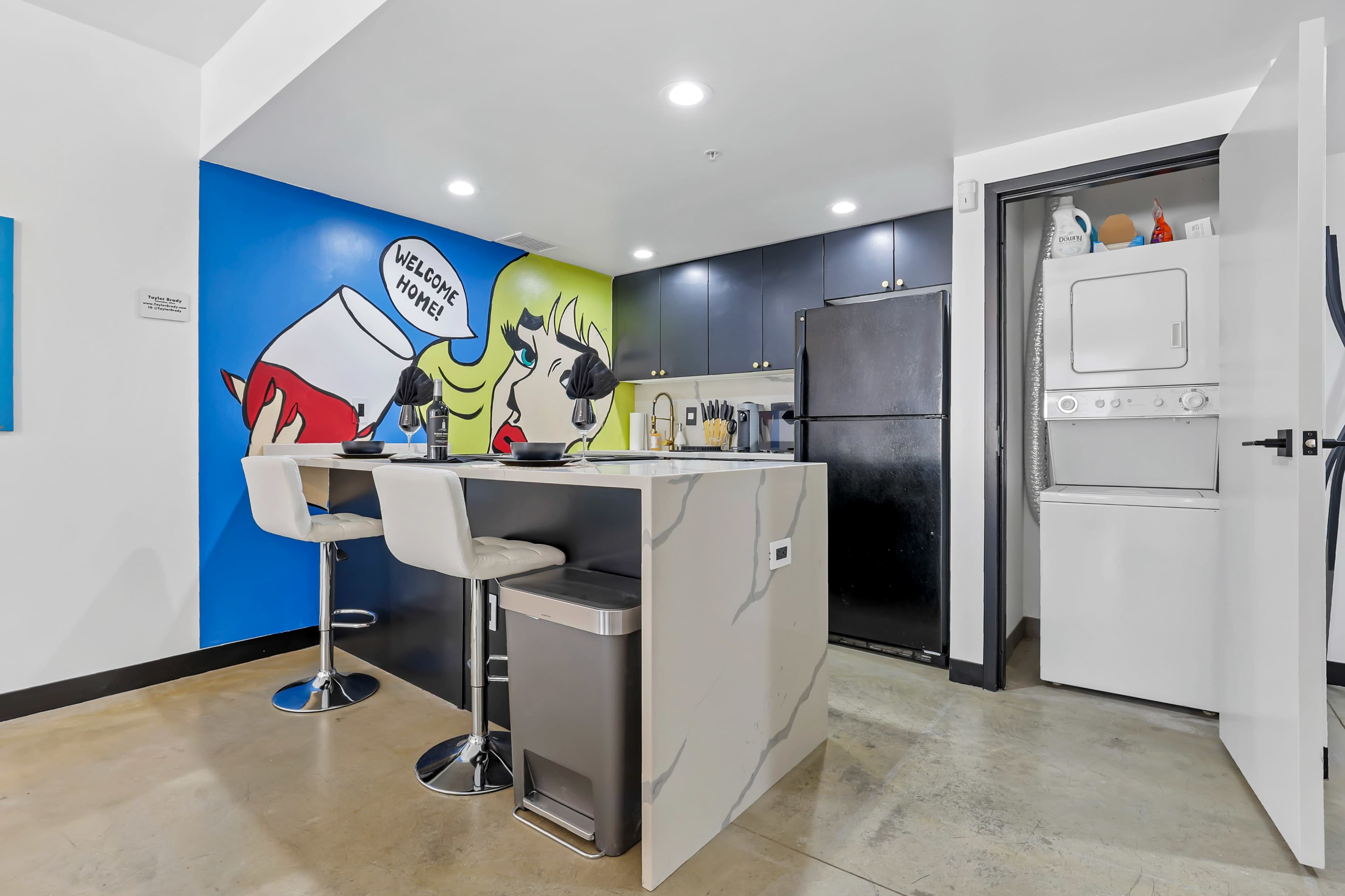 A modern kitchen with a colorful mural, featuring a woman holding a beverage, and a compact laundry area next to black appliances.