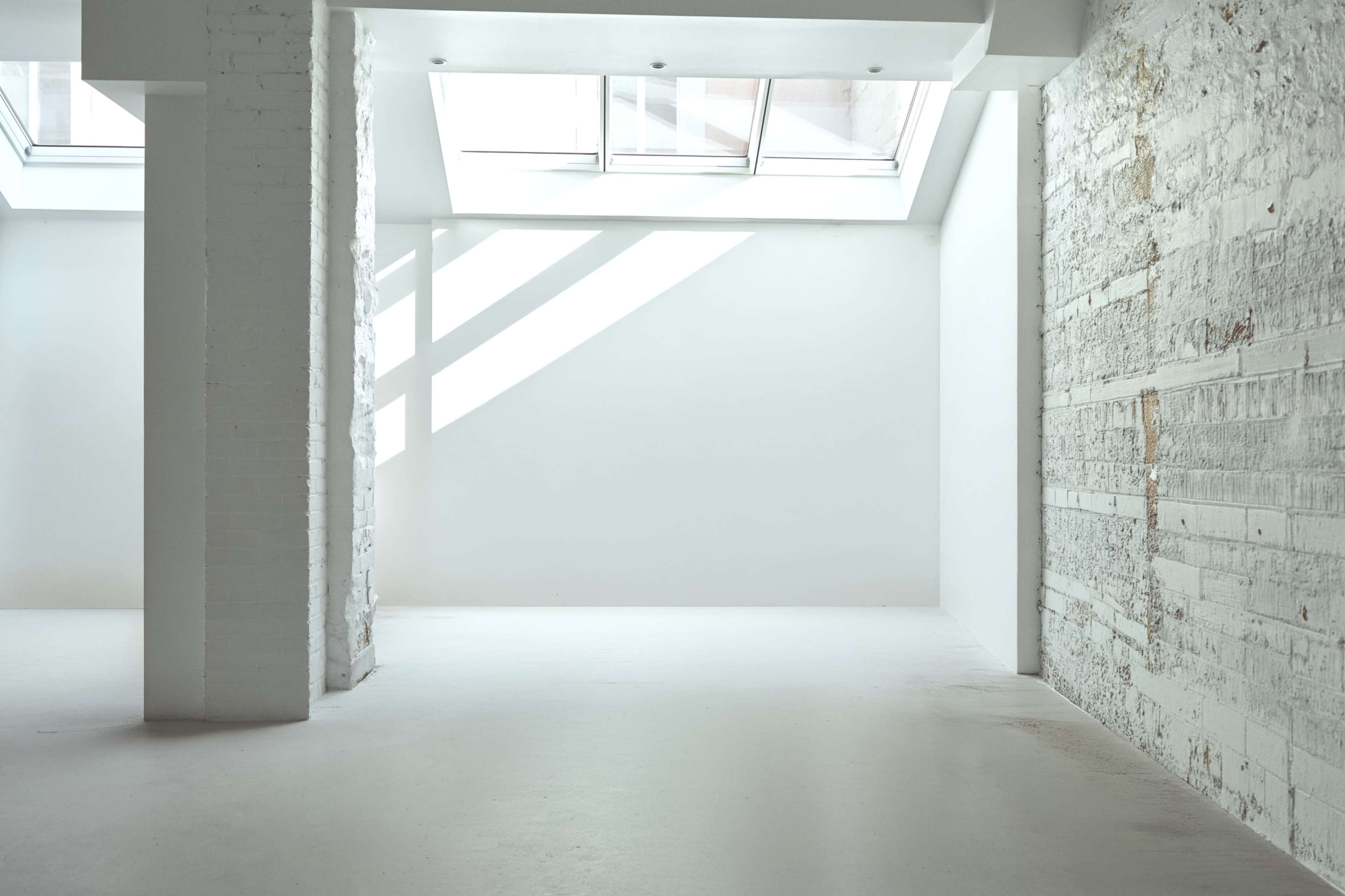 The image shows an empty room with bright white walls and large skylights allowing natural light to filter in.