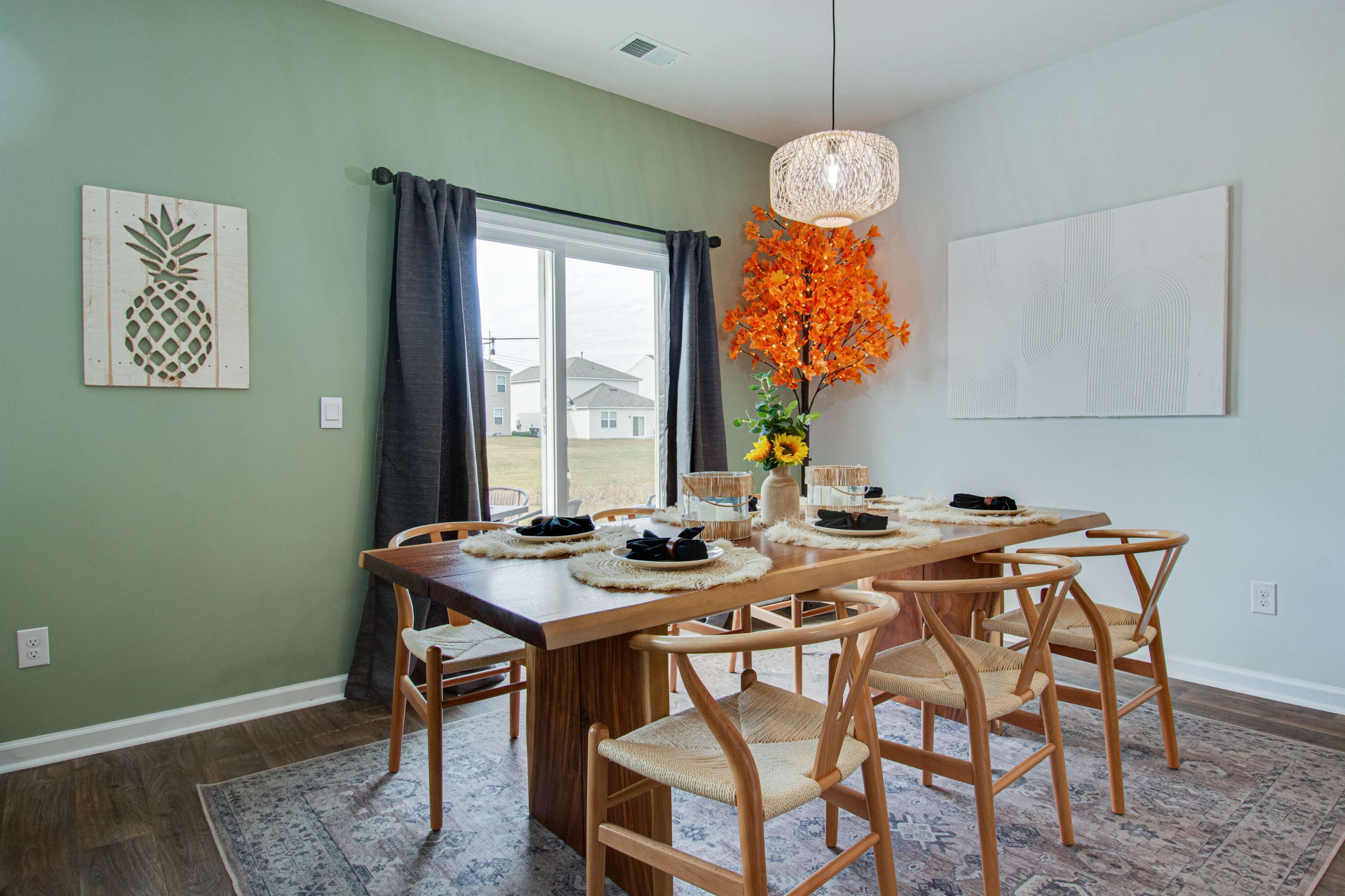 A dining room features a wooden table surrounded by chairs, a vase of orange flowers, and a large window with curtains.
