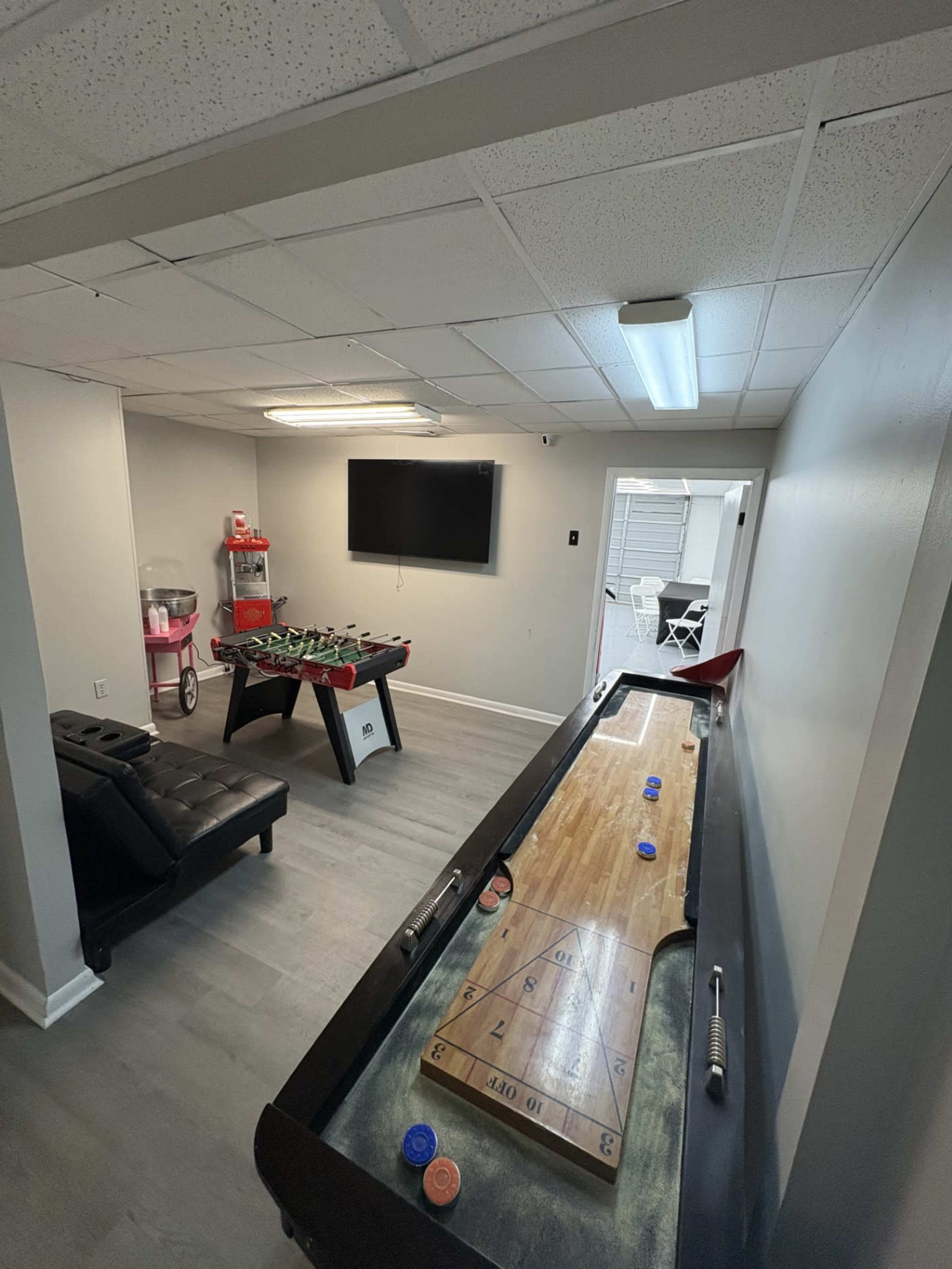 The image shows a recreational room featuring a shuffleboard table, a foosball table, a black sofa, and a wall-mounted television.