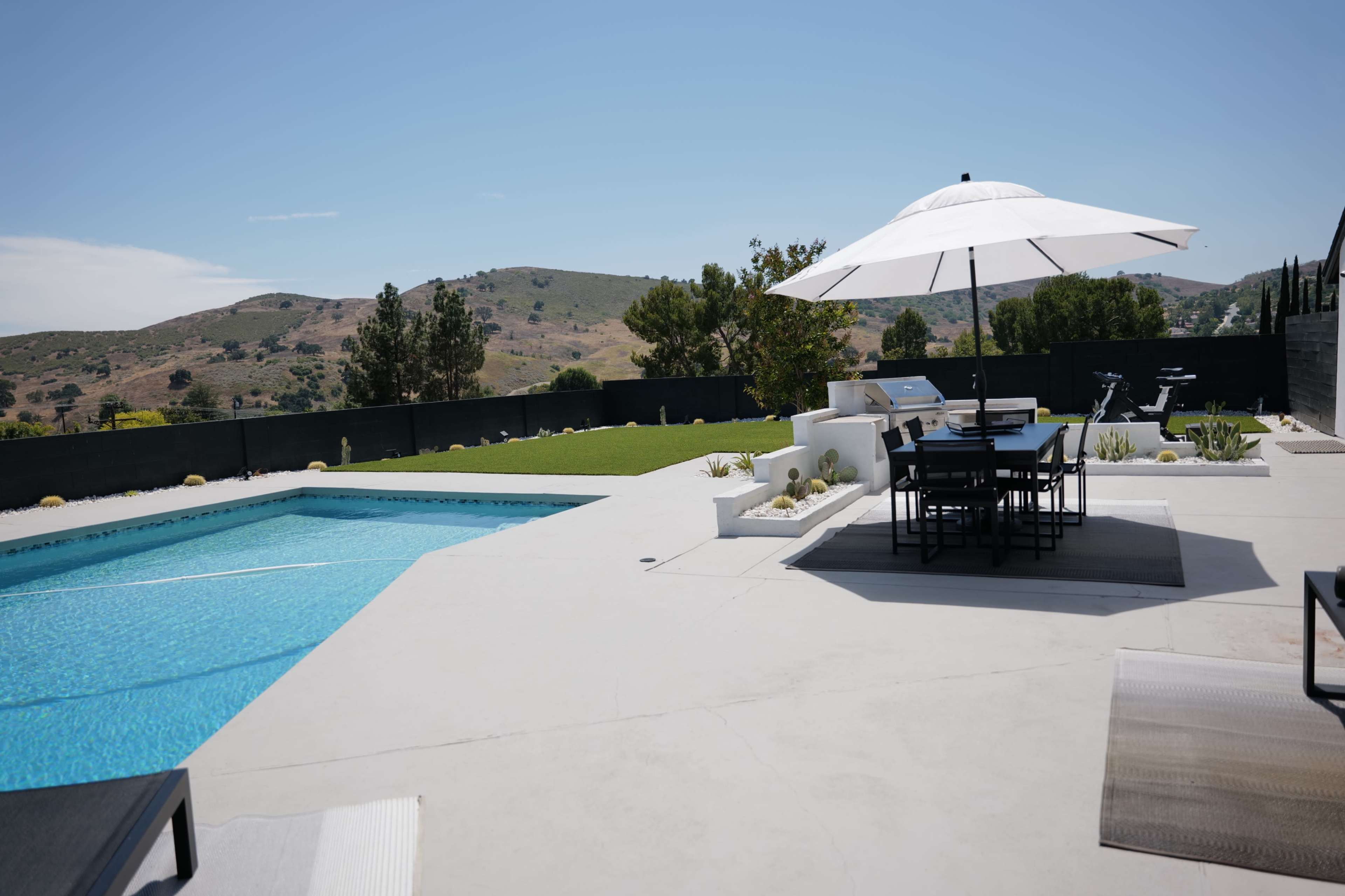 The image shows a modern backyard with a swimming pool, a dining table under a large umbrella, and a view of rolling hills in the background.