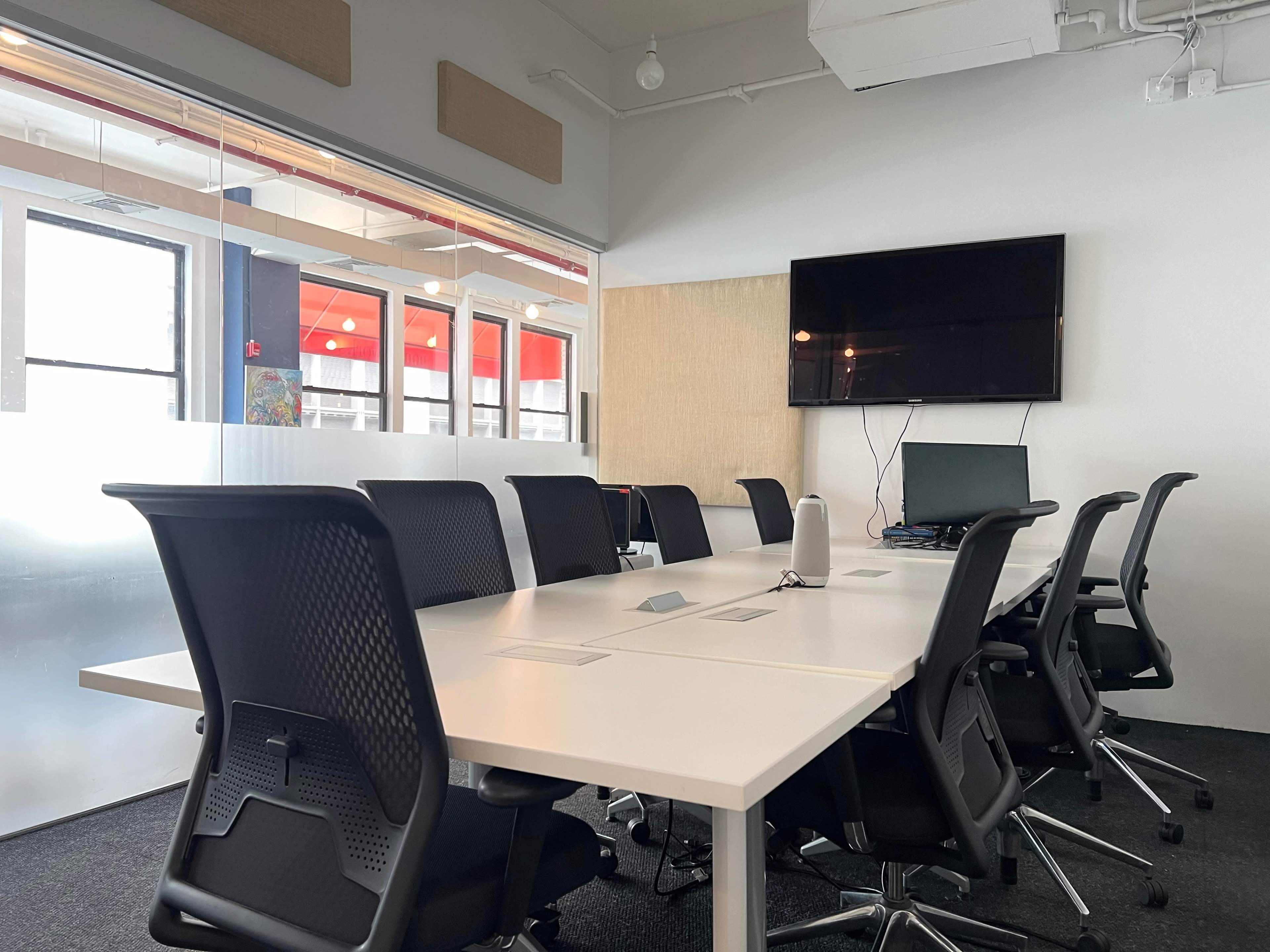 A modern conference room features a large white table surrounded by black office chairs, with a wall-mounted TV and computer setup in the background.