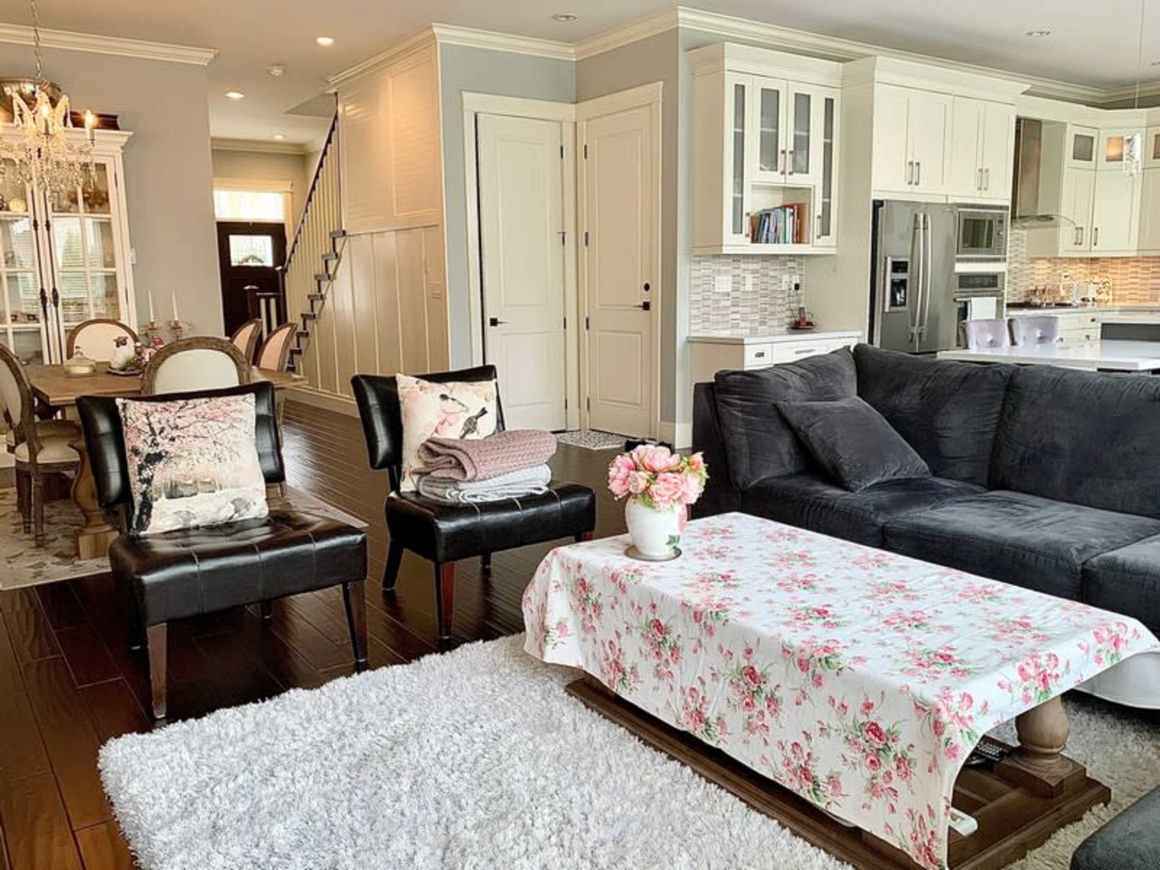 The living room features a dark sectional sofa, two black chairs with floral pillows, a coffee table draped with a floral cloth, and a white rug.