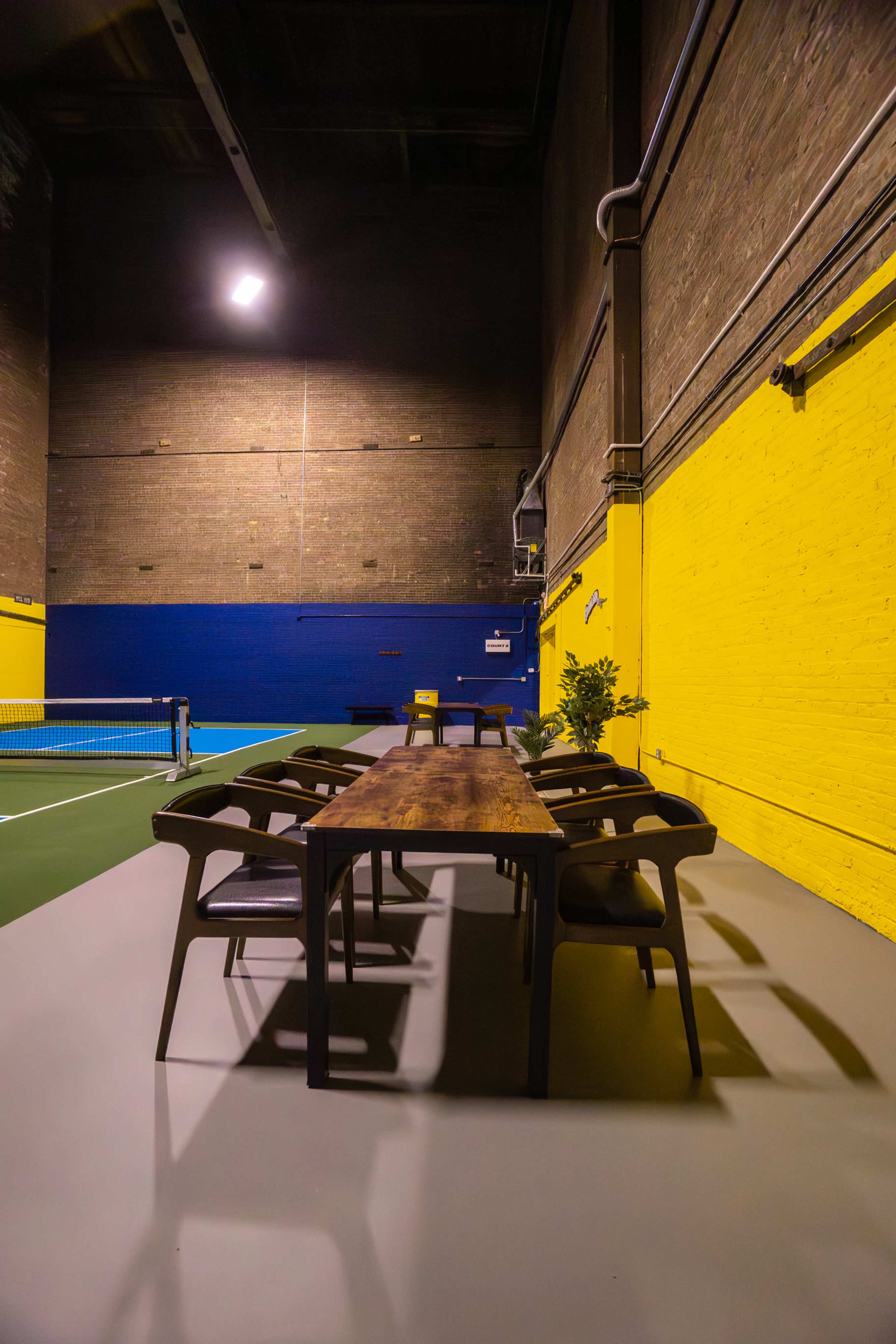 A long wooden table with chairs is set in an industrial space featuring a vibrant yellow and blue wall, adjacent to a tennis court.