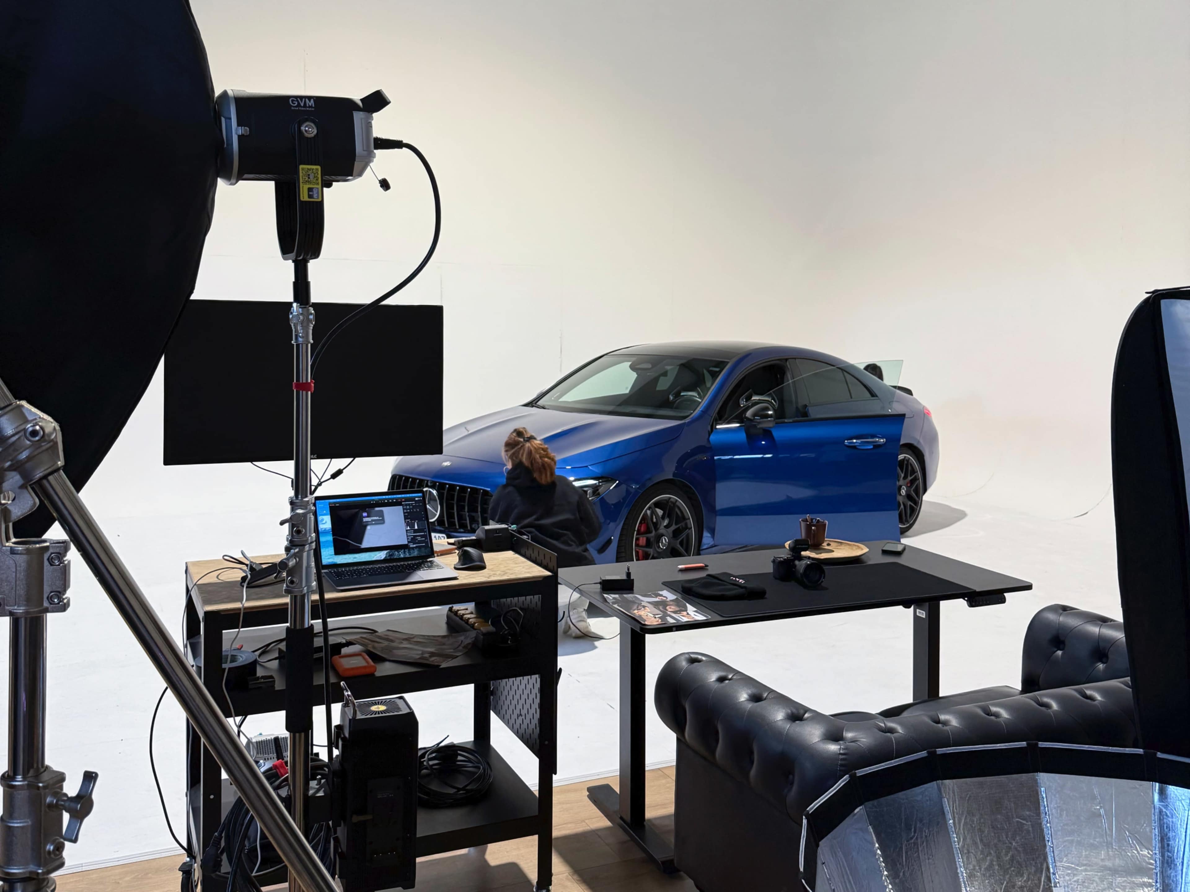 A blue luxury car is positioned in a well-lit studio next to a black desk with a laptop and various items.