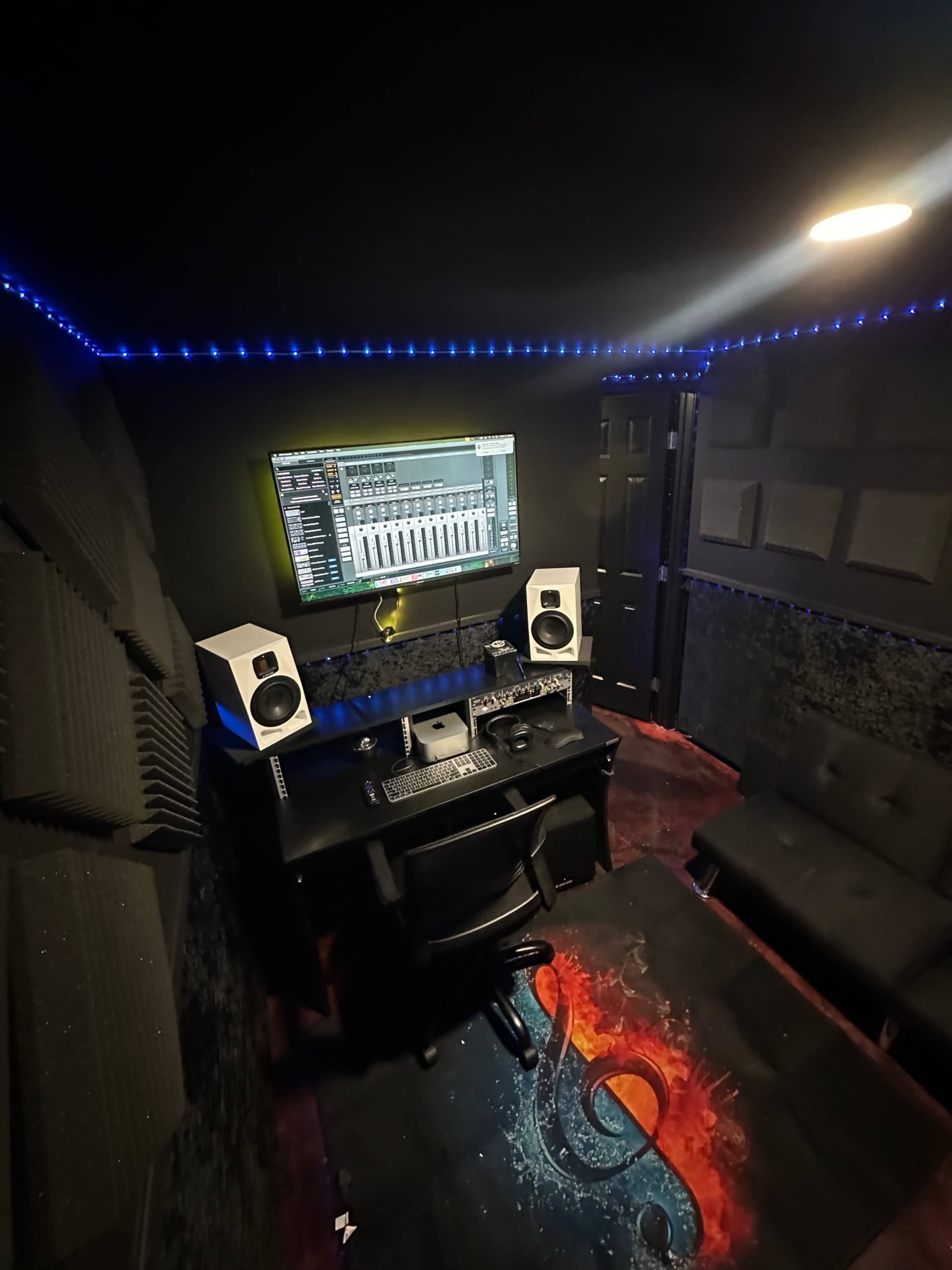 The image shows a small music production studio with soundproof panels, a computer monitor displaying audio software, studio monitors on a desk, and a sofa in a dimly lit room accented by blue LED lights.