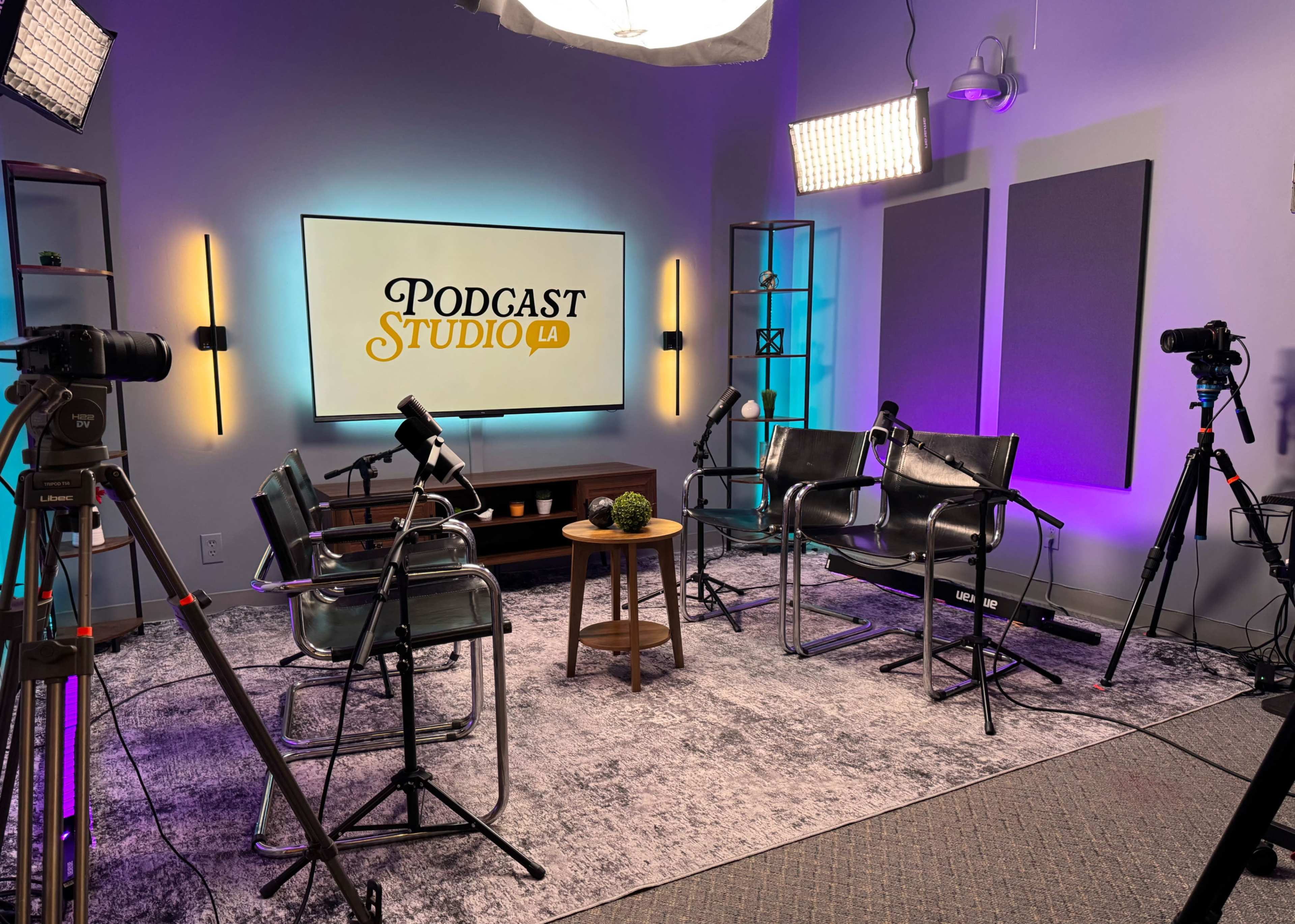The image shows a podcast studio with two chairs, a small wooden table, and multiple cameras set up in front of a large screen displaying the text "PODCAST STUDIO LA."