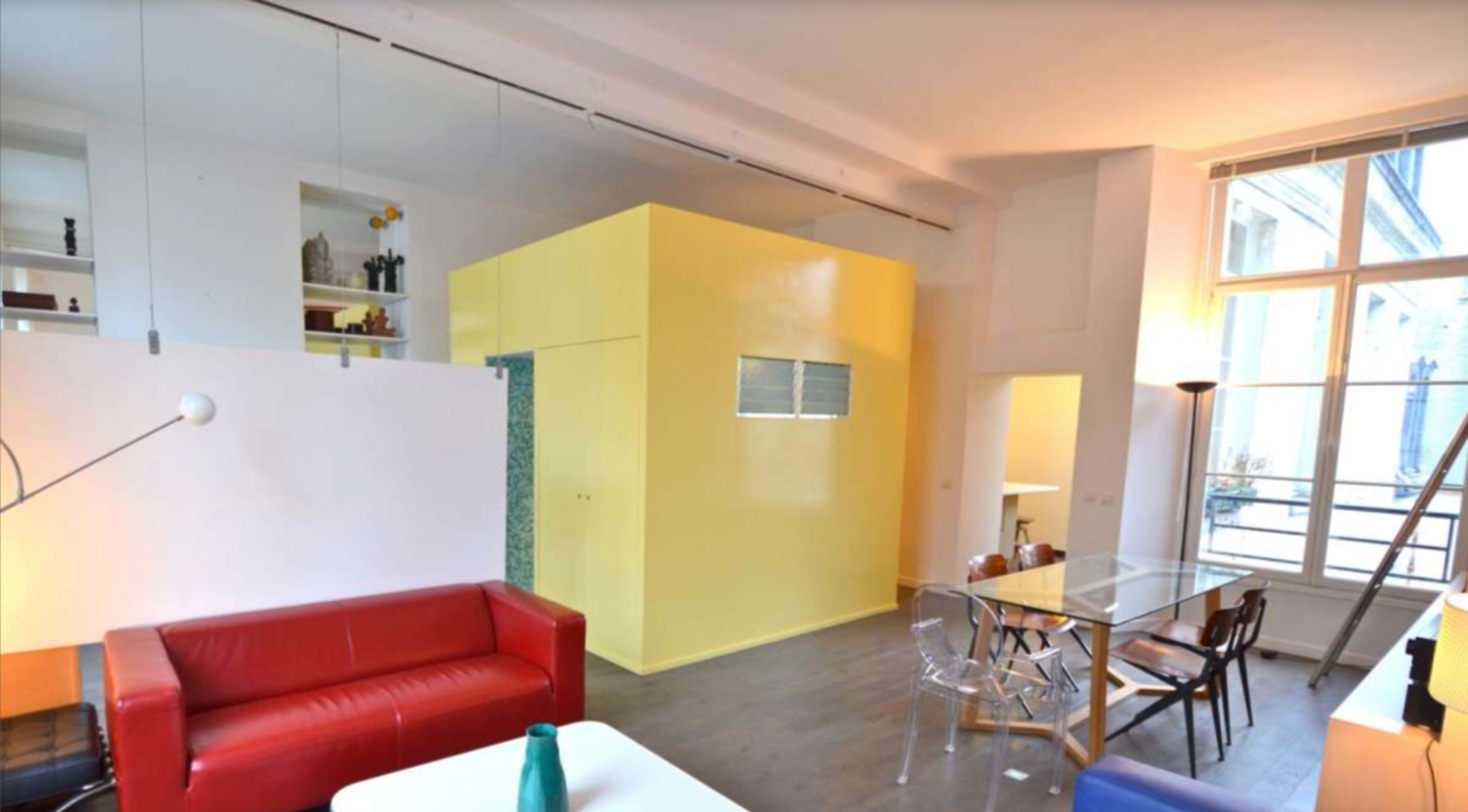 A spacious interior with modern furniture, featuring a bold yellow room divider and a dining area with a transparent table and wooden chairs.