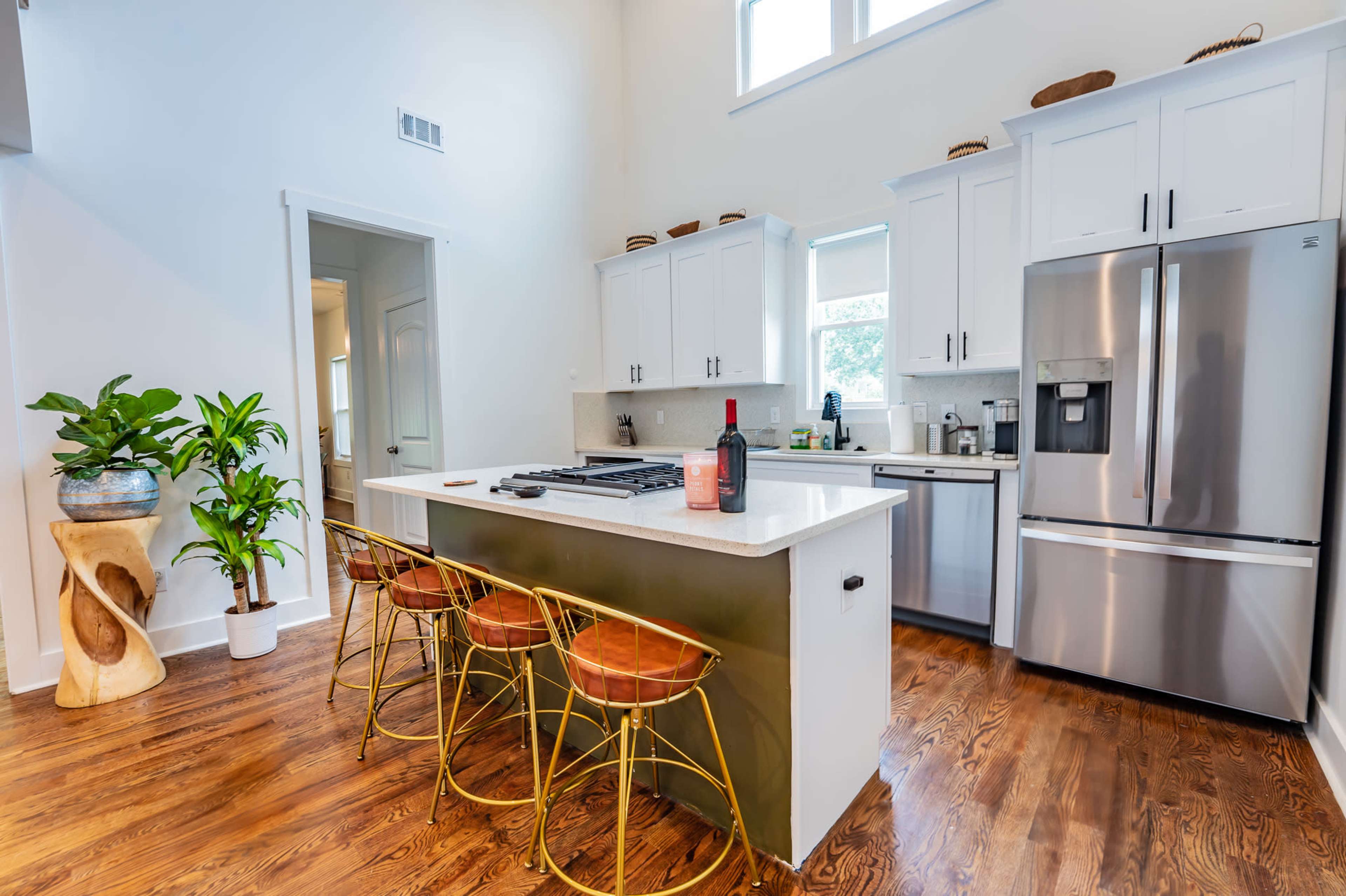 A modern kitchen features a central island with three gold bar stools, stainless steel appliances, and a large window providing natural light.