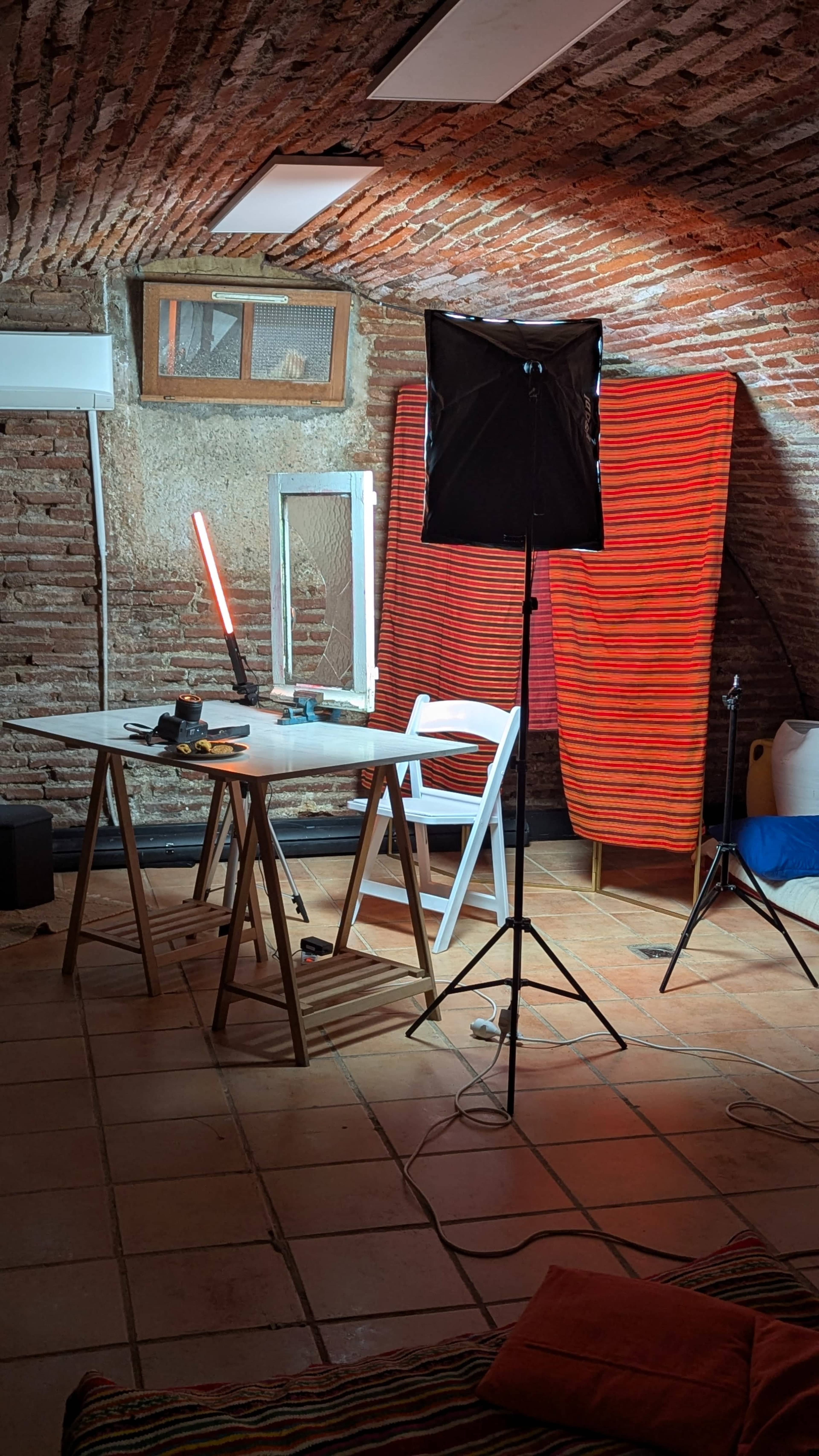 The scene features a cozy attic room with exposed brick walls, a table set up with various equipment, and a backdrop made of colorful fabric.