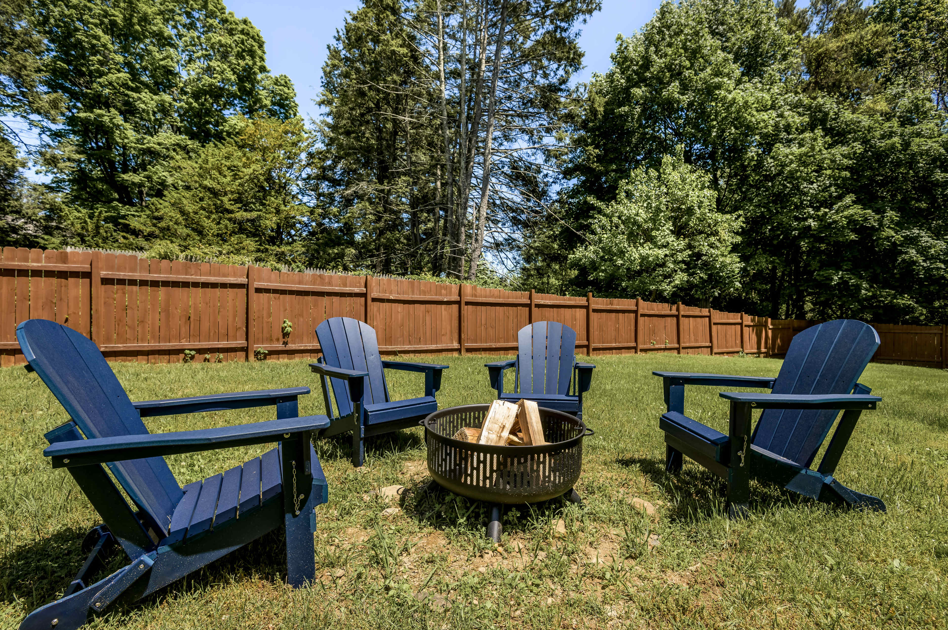 A set of four blue Adirondack chairs surrounds a fire pit in a grassy backyard with a wooden fence and trees in the background.