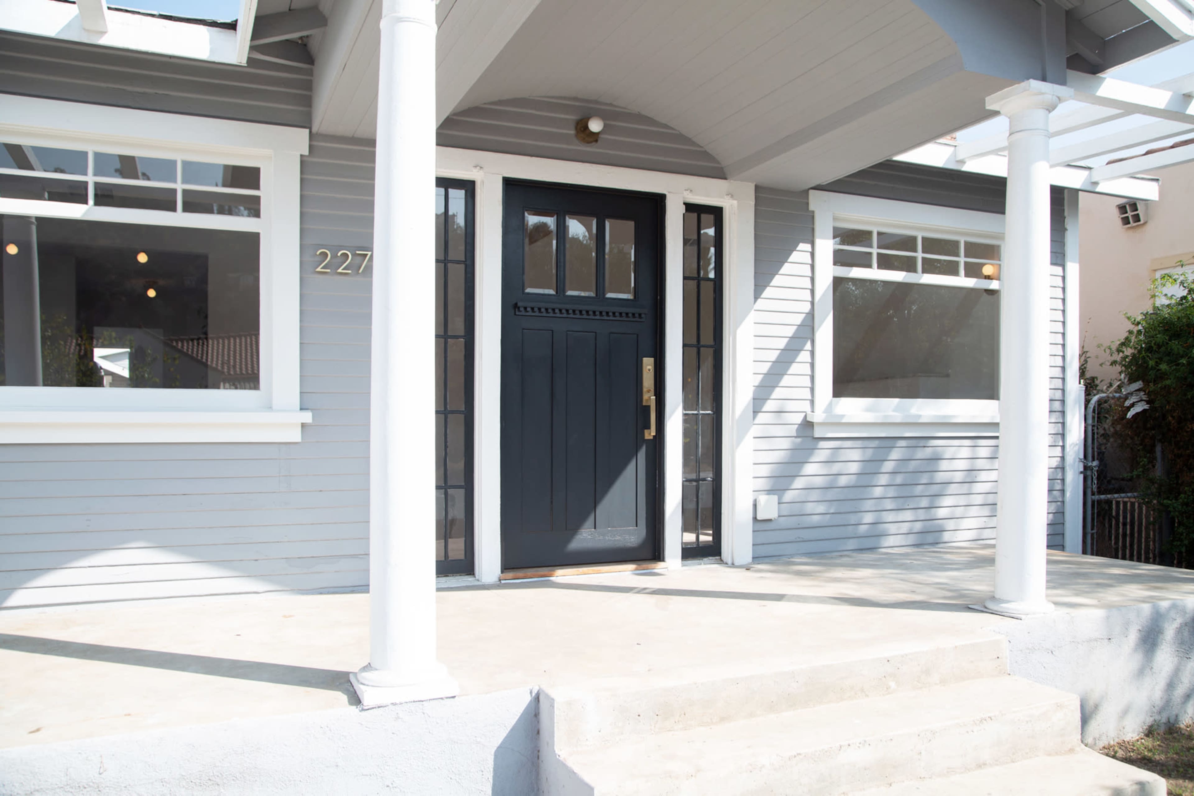 The image shows a front entrance of a grey house featuring a black door, white pillars, and a concrete porch.