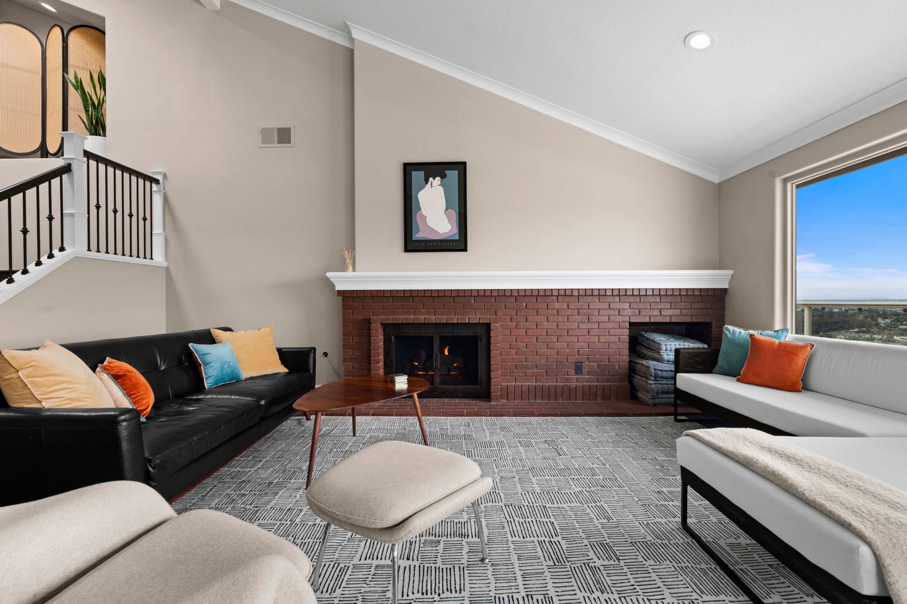 A modern living room features a black leather sofa, a white sectional, a coffee table, and a brick fireplace, with large windows offering a view outside.