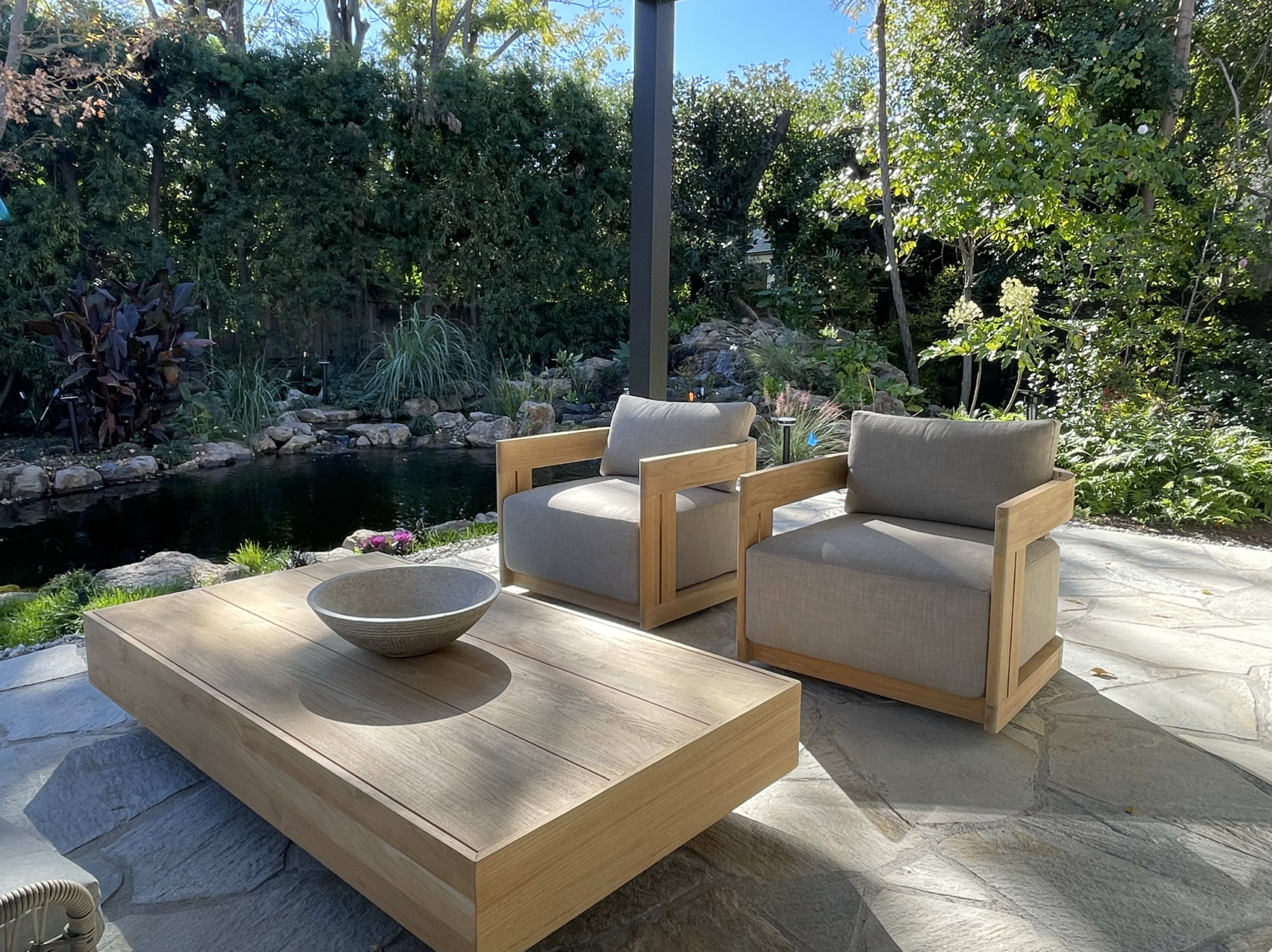 The image shows two wooden armchairs with gray cushions positioned beside a stone coffee table in a landscaped garden with a pond.