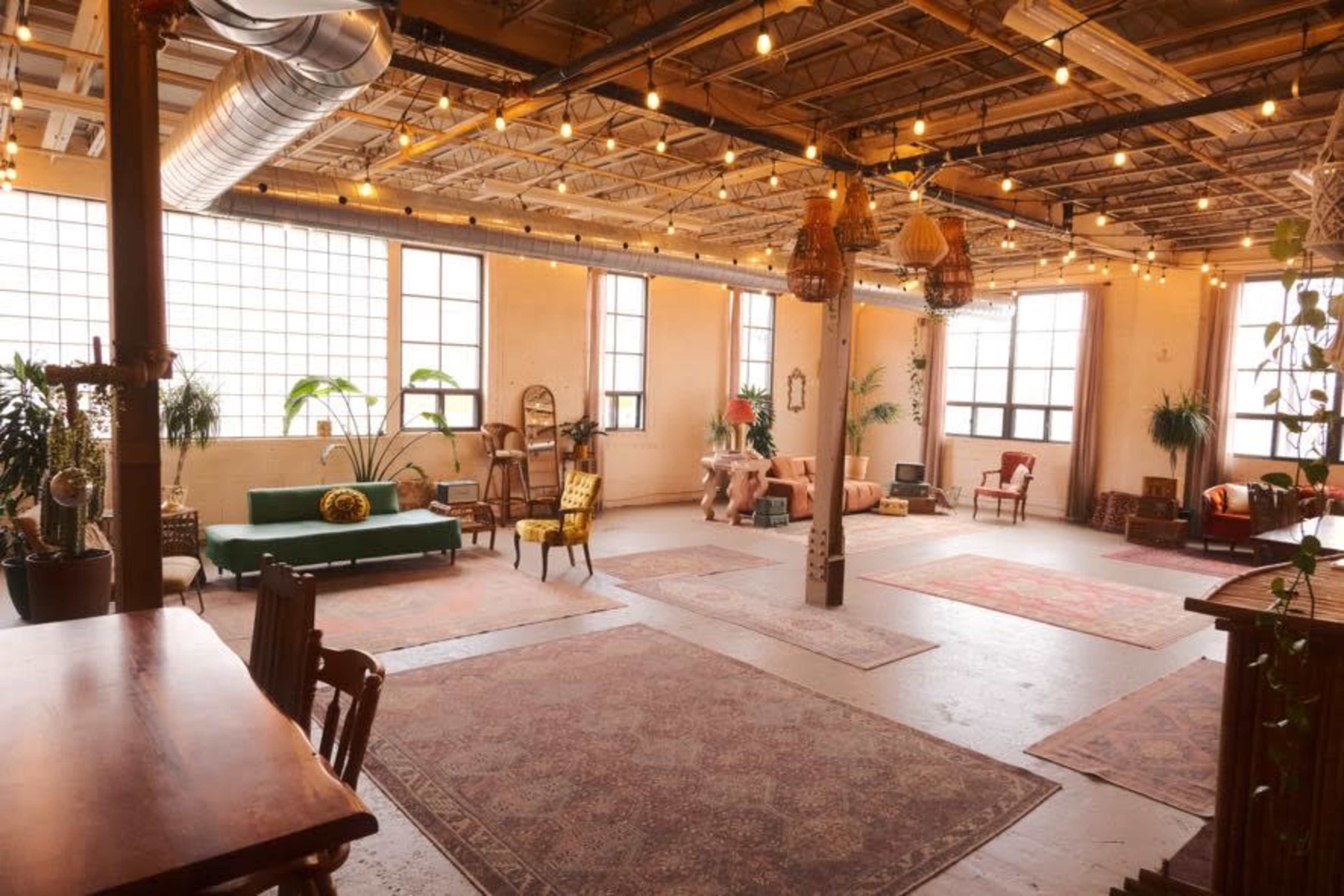 The image shows an open, well-lit industrial space with multiple seating areas, decorative rugs, and hanging string lights.