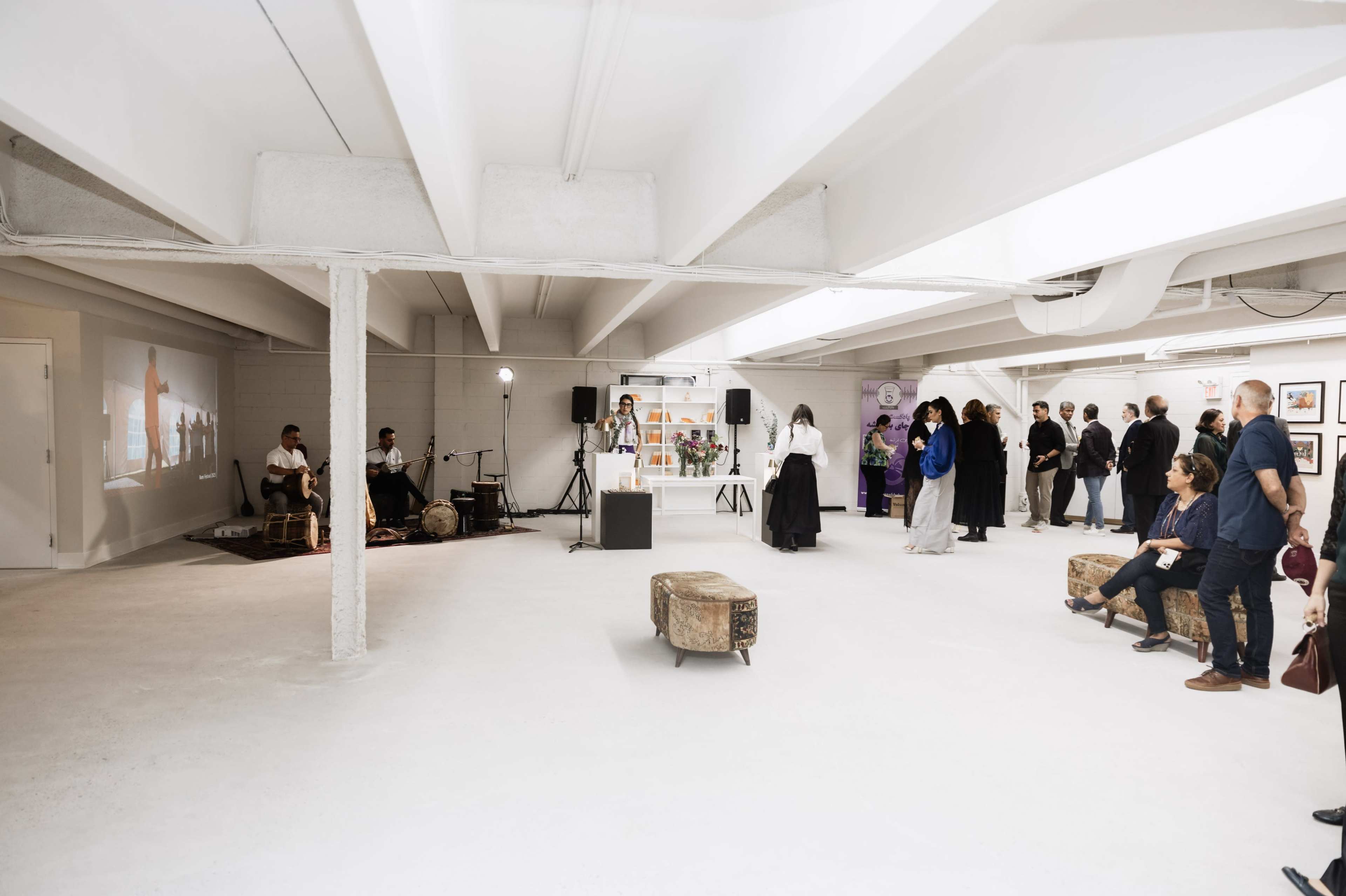 A gallery space features a performance area with musicians, a projection on the wall, and an audience engaging with an art exhibit.