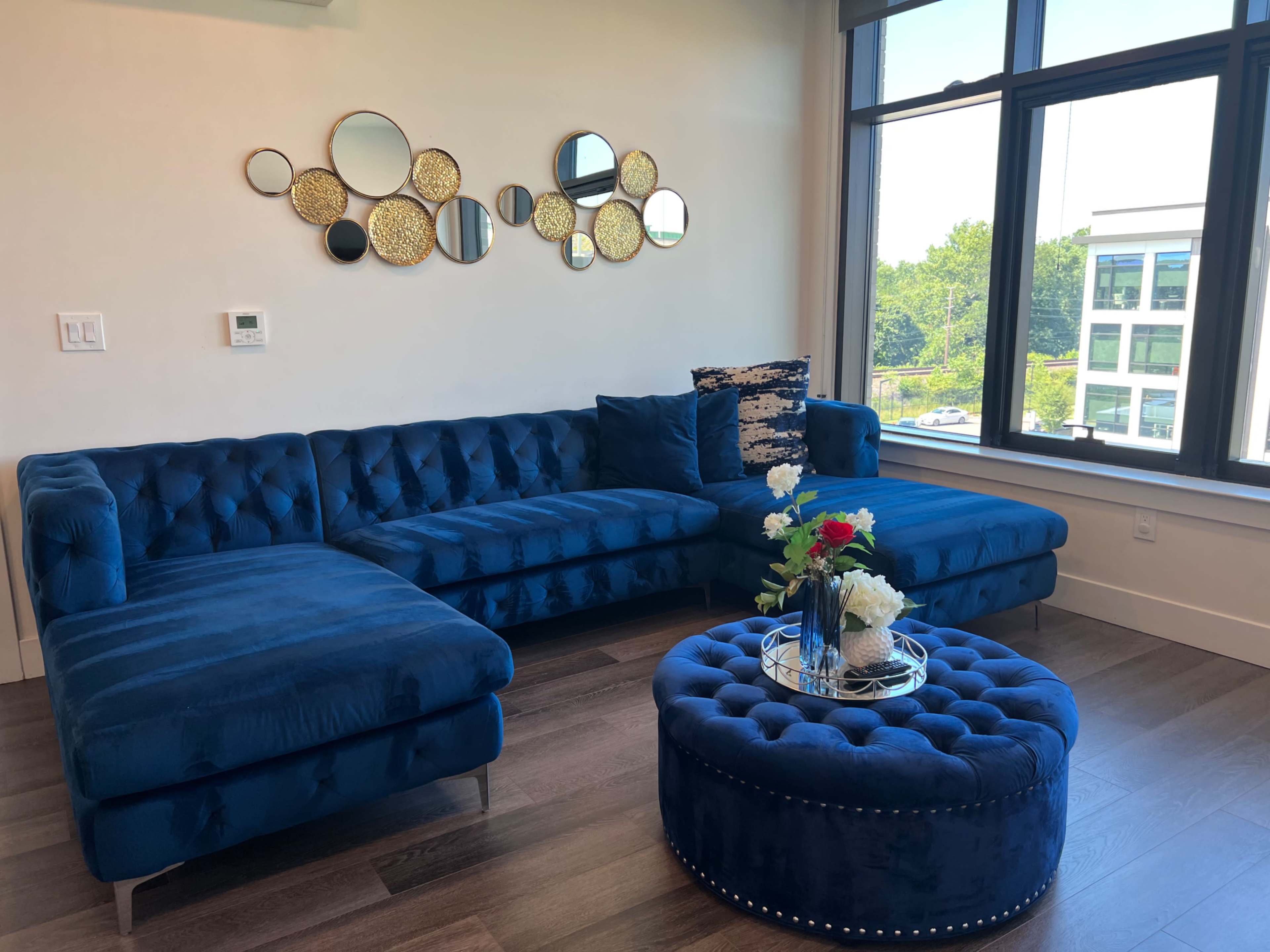 A modern living room featuring a large blue tufted sectional sofa, a round ottoman, and decorative mirrors on the wall.