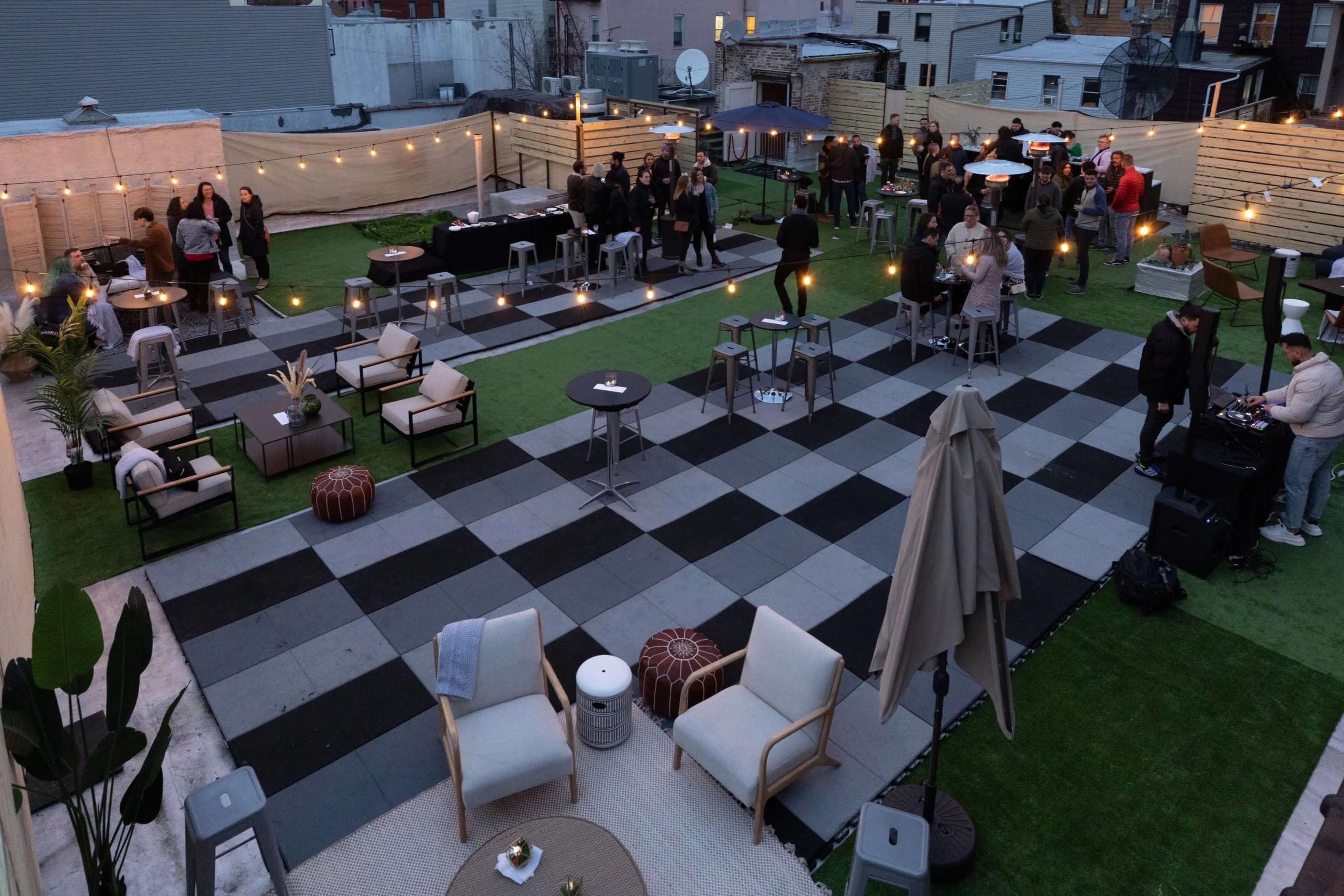 The image shows a rooftop event space with a checkerboard pattern of black and white tiles, seating areas, and string lights illuminating a gathering of people.