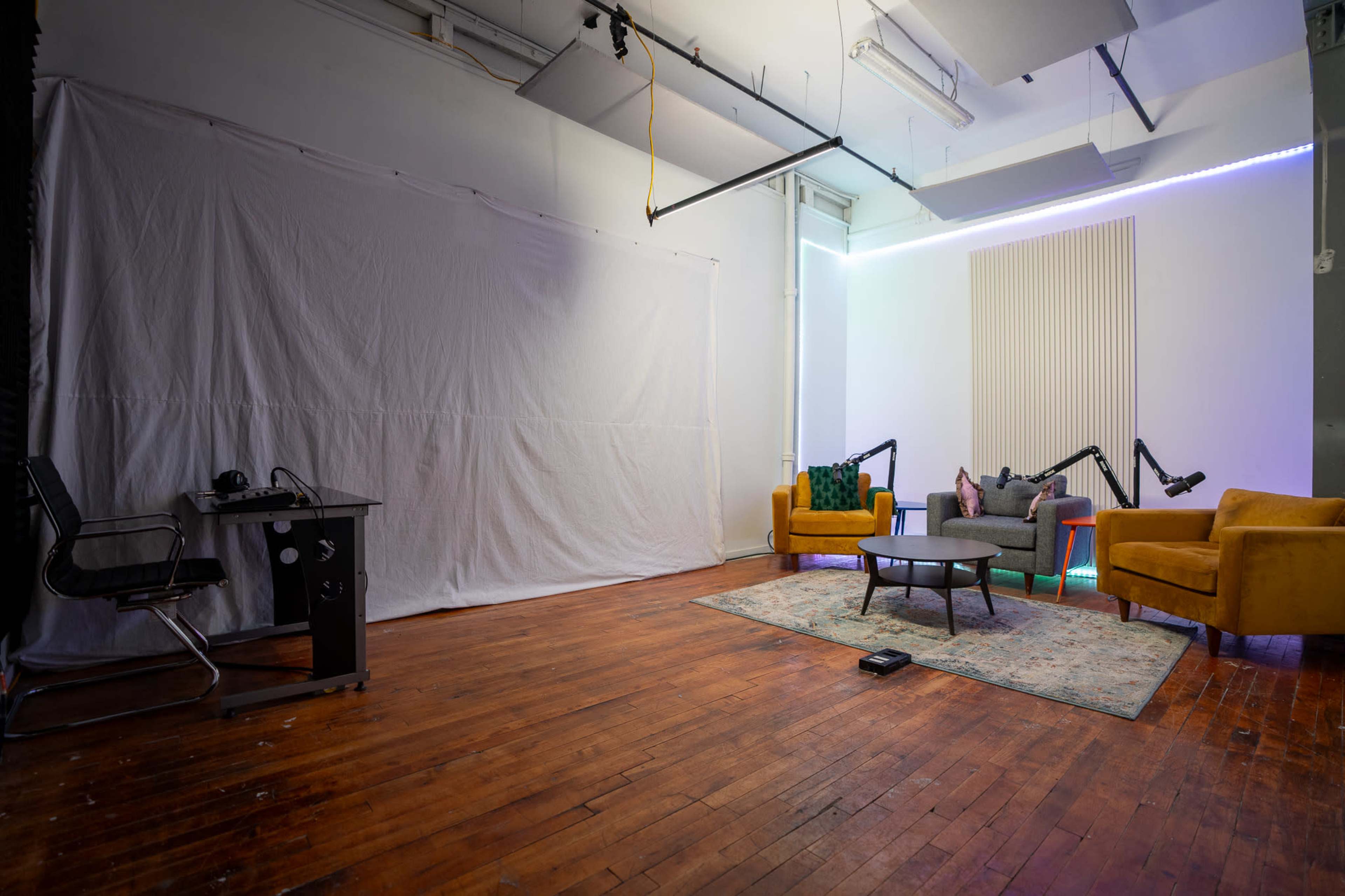 The image shows an empty studio space with a white backdrop, a wooden floor, a desk with a chair, and two yellow armchairs arranged around a small rug.