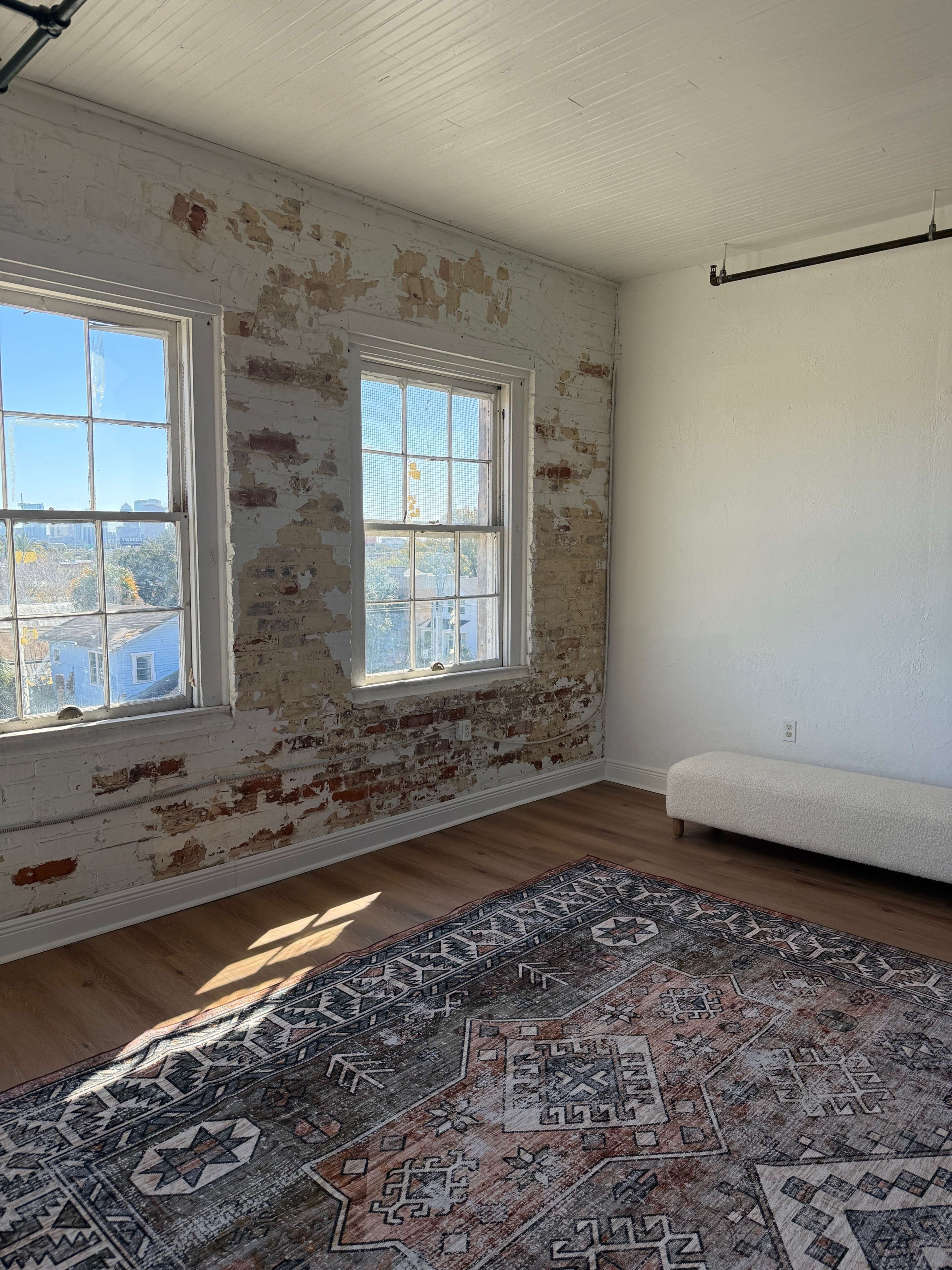 The image shows a room with exposed brick walls, two large windows, and a patterned rug on a wooden floor.