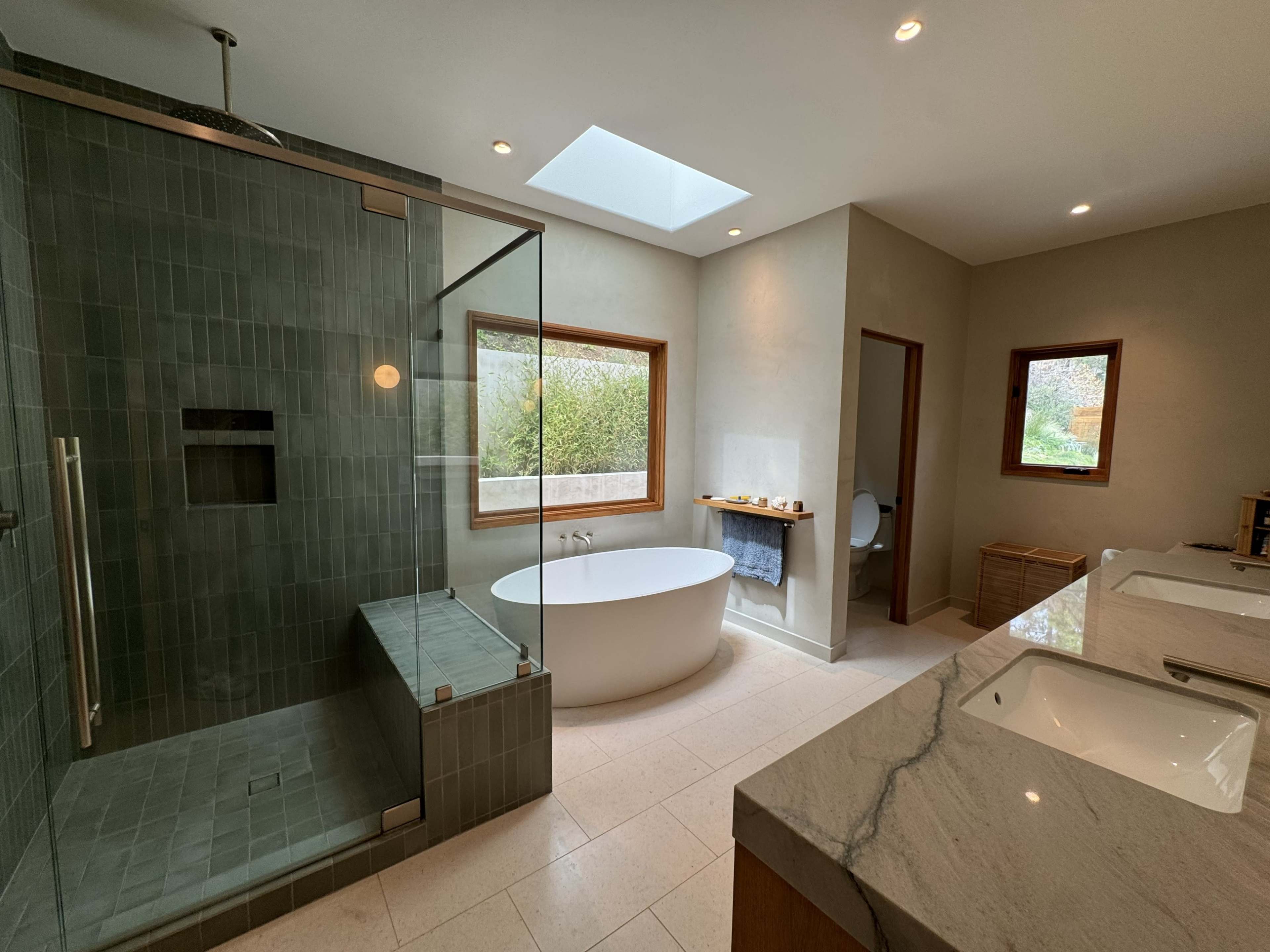 The image shows a modern bathroom featuring a walk-in shower, a freestanding bathtub, dual sinks with marble countertops, and large windows providing natural light.