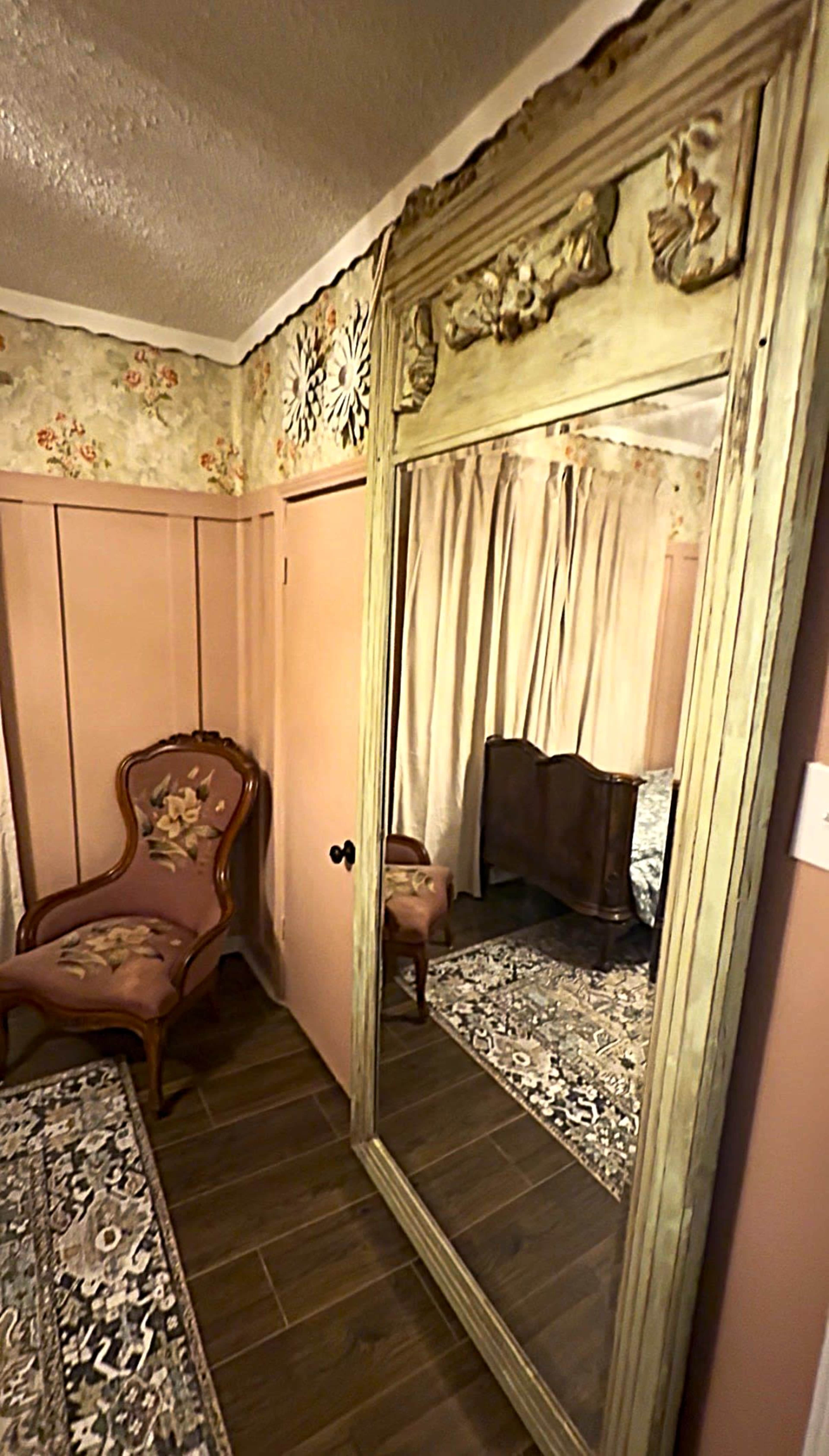 The image shows a vintage-style room featuring a large ornate mirror, a patterned rug, and a floral wallpaper with a wooden chair and bed in the background.