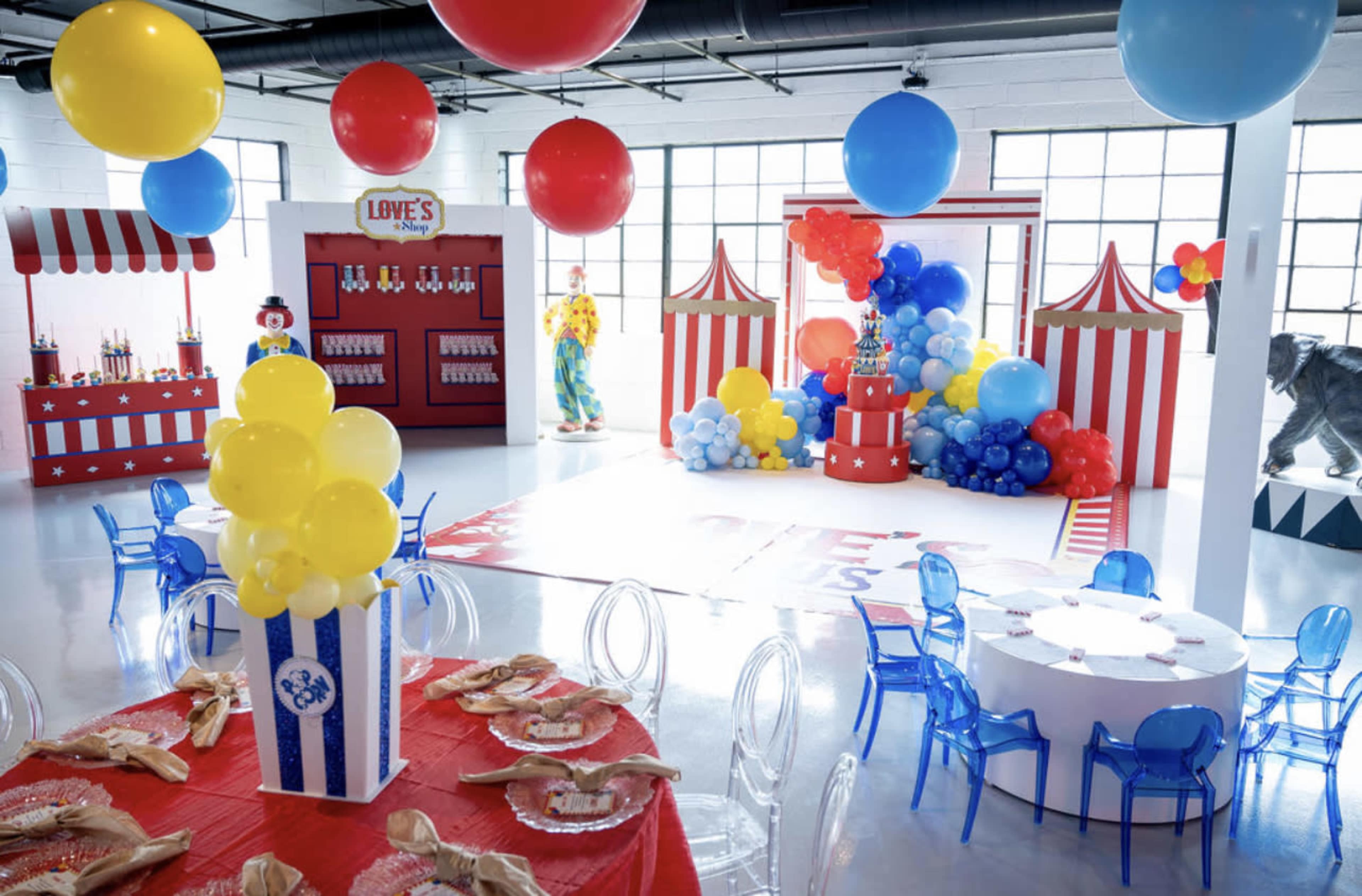 A colorful party venue decorated with large balloons, a candy stand, and a circus-themed setup featuring tables, chairs, and a festive cake display.