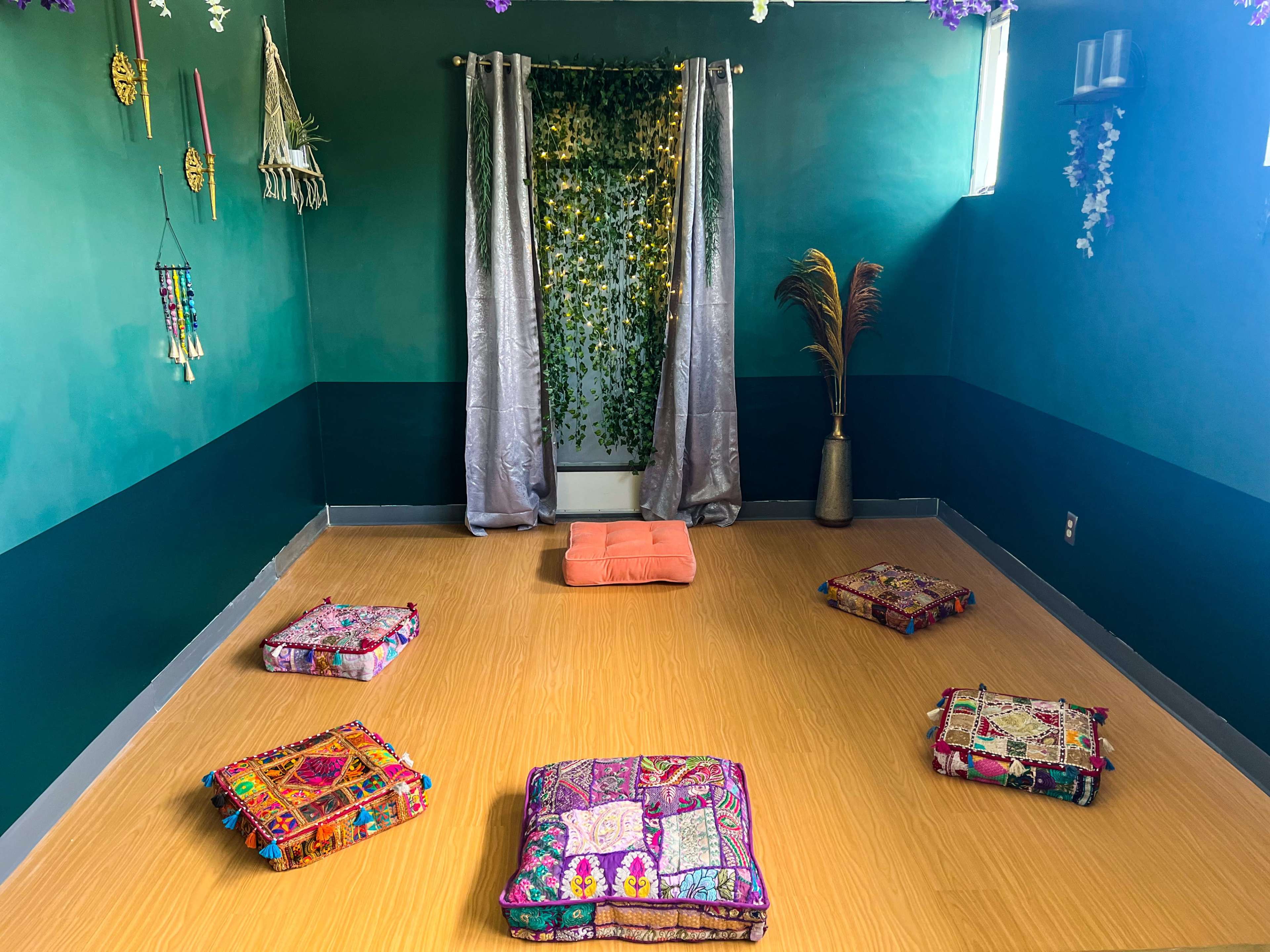 The scene shows a tranquil room with a wooden floor, decorated with colorful cushions arranged in a circle, a backdrop of green vines, and a pair of curtains framing a light source.