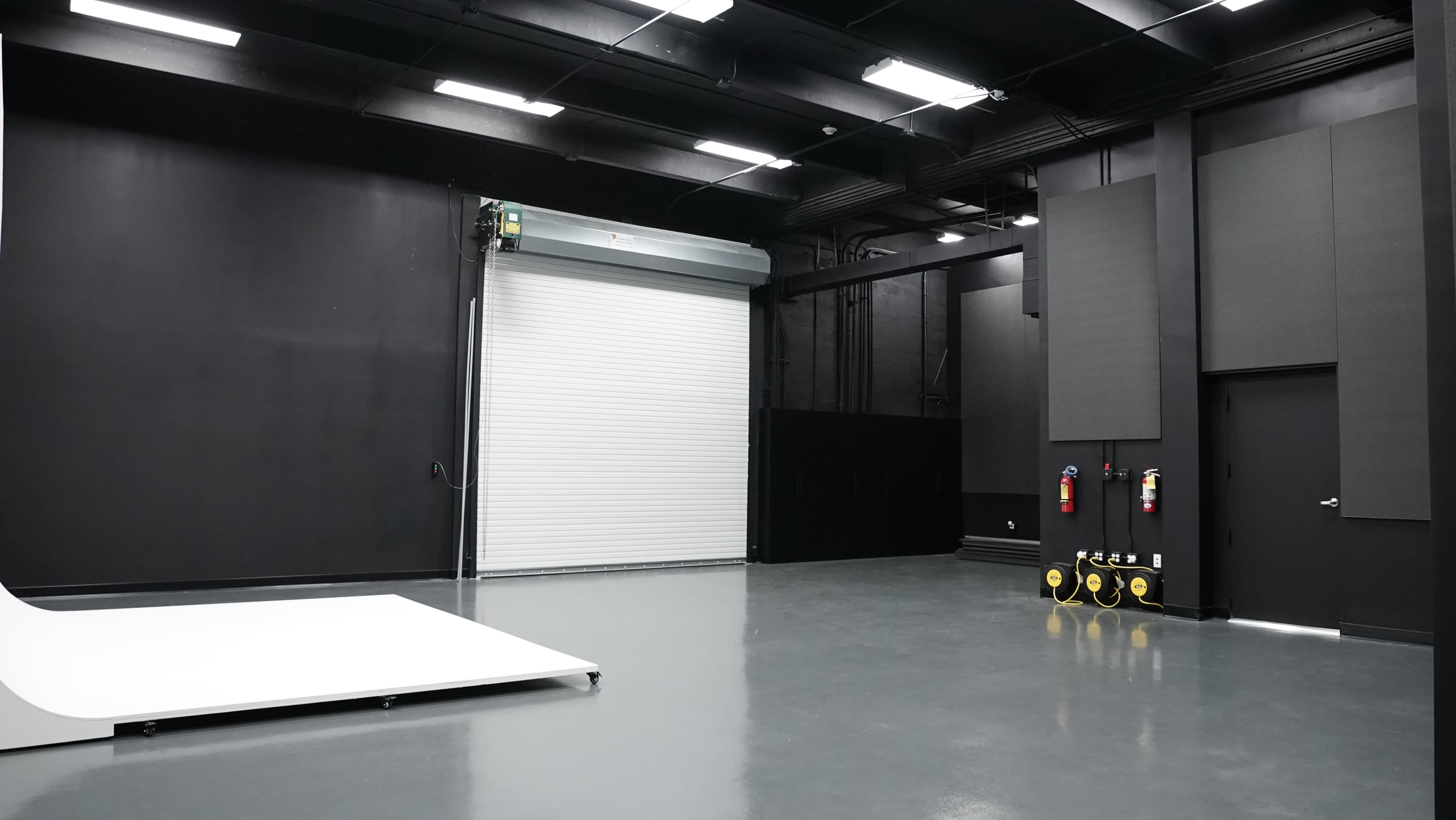 The image shows an empty studio space with a white stage area, a roll-up door, and safety equipment on the wall.