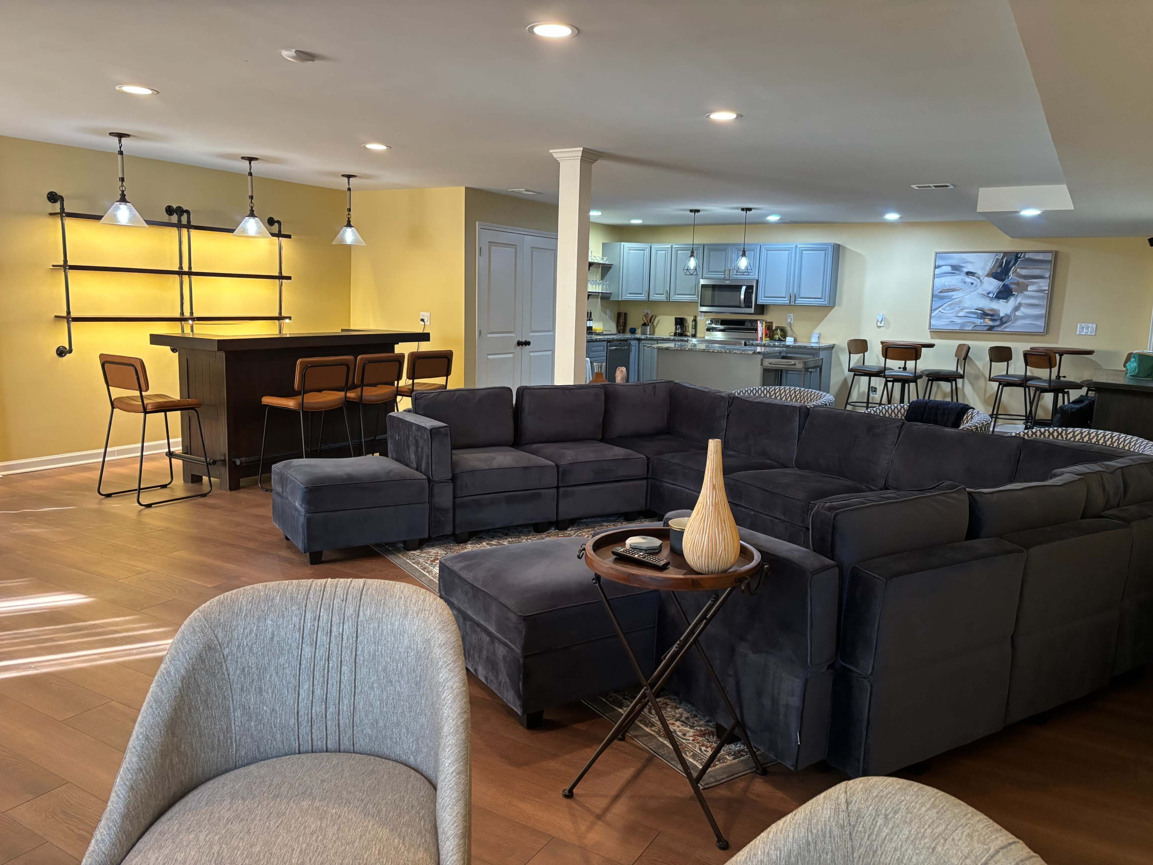 The image shows a modern living area with a large sectional sofa, a small side table, and a bar area in the background, featuring a fully equipped kitchen.