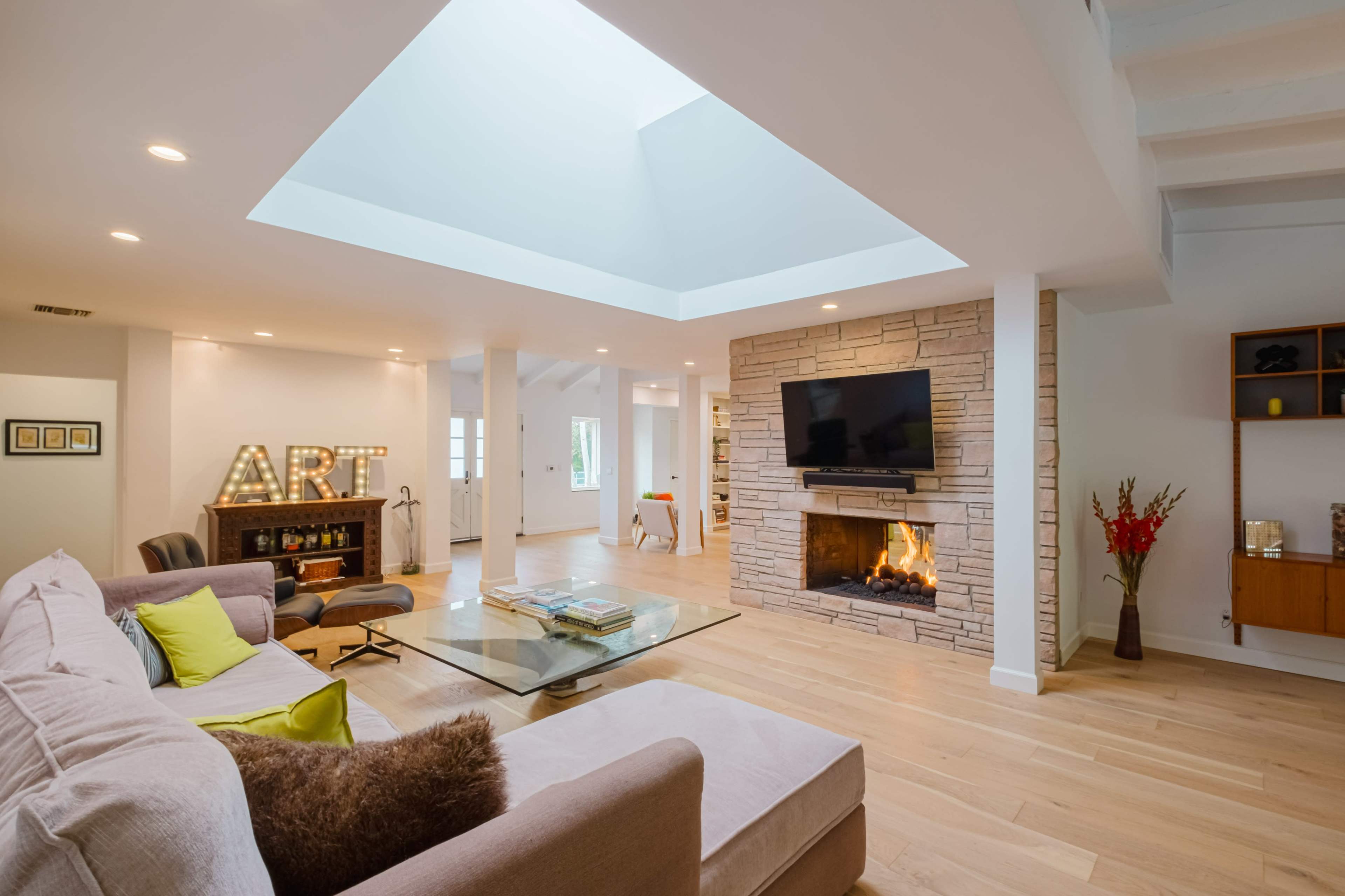 A spacious living room features a stone fireplace, a skylight, and a modern sofa arranged around a glass coffee table.