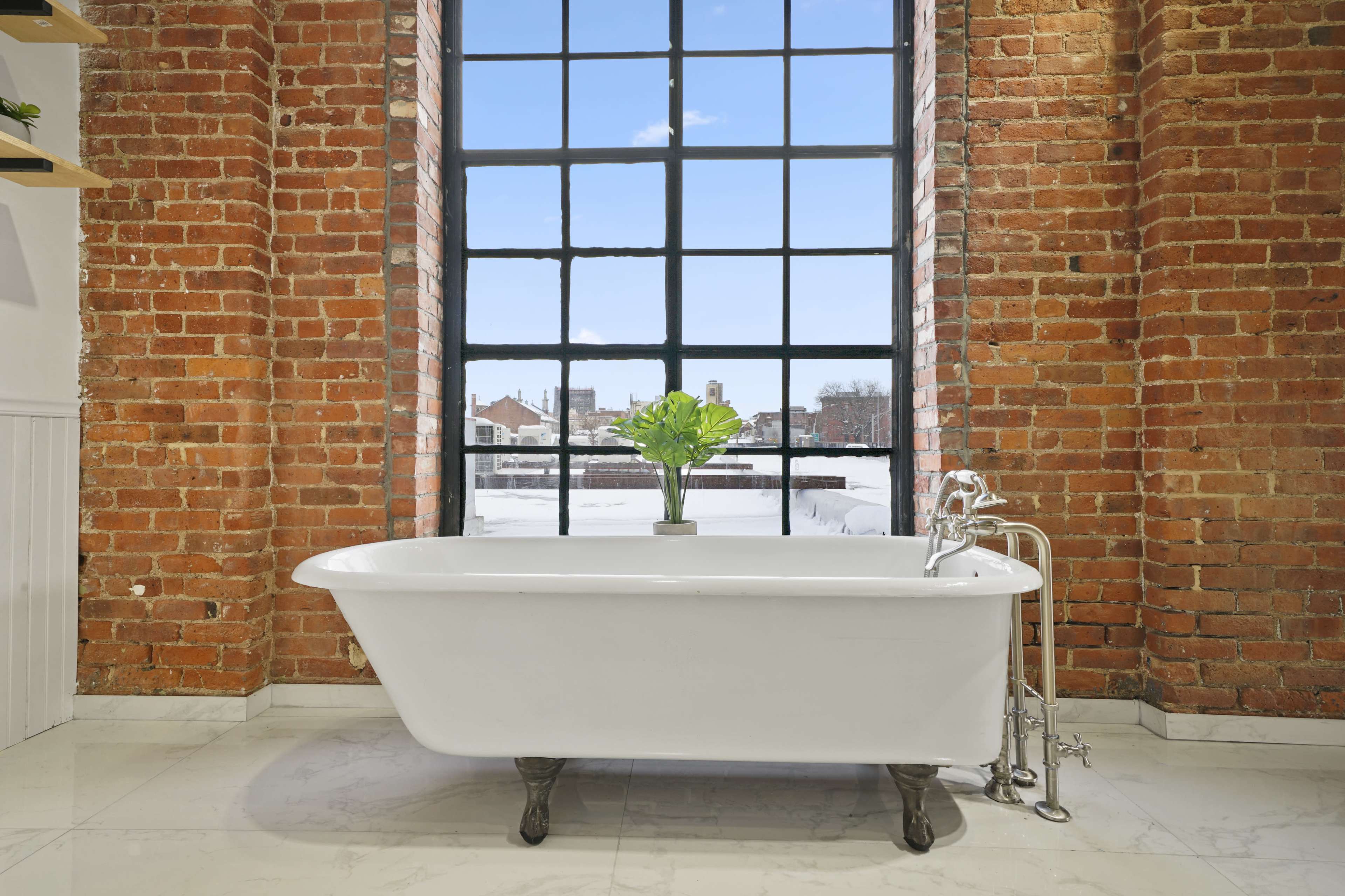 A white clawfoot bathtub is positioned in front of a large industrial-style window with a view of an outside landscape, surrounded by exposed brick walls.