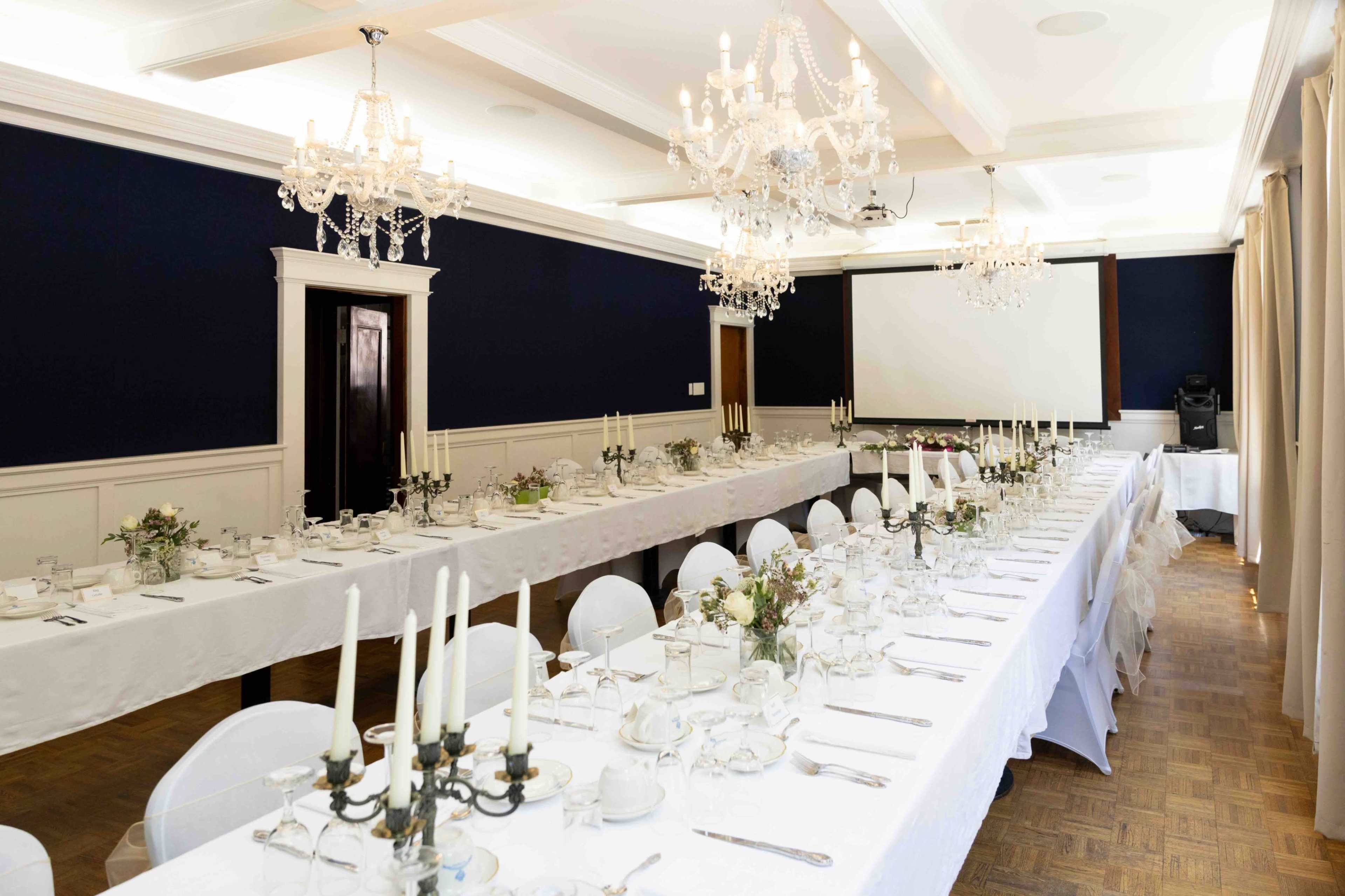 A large dining room features a long table set with white tablecloths, candles, and elegant dishware, illuminated by several chandeliers.