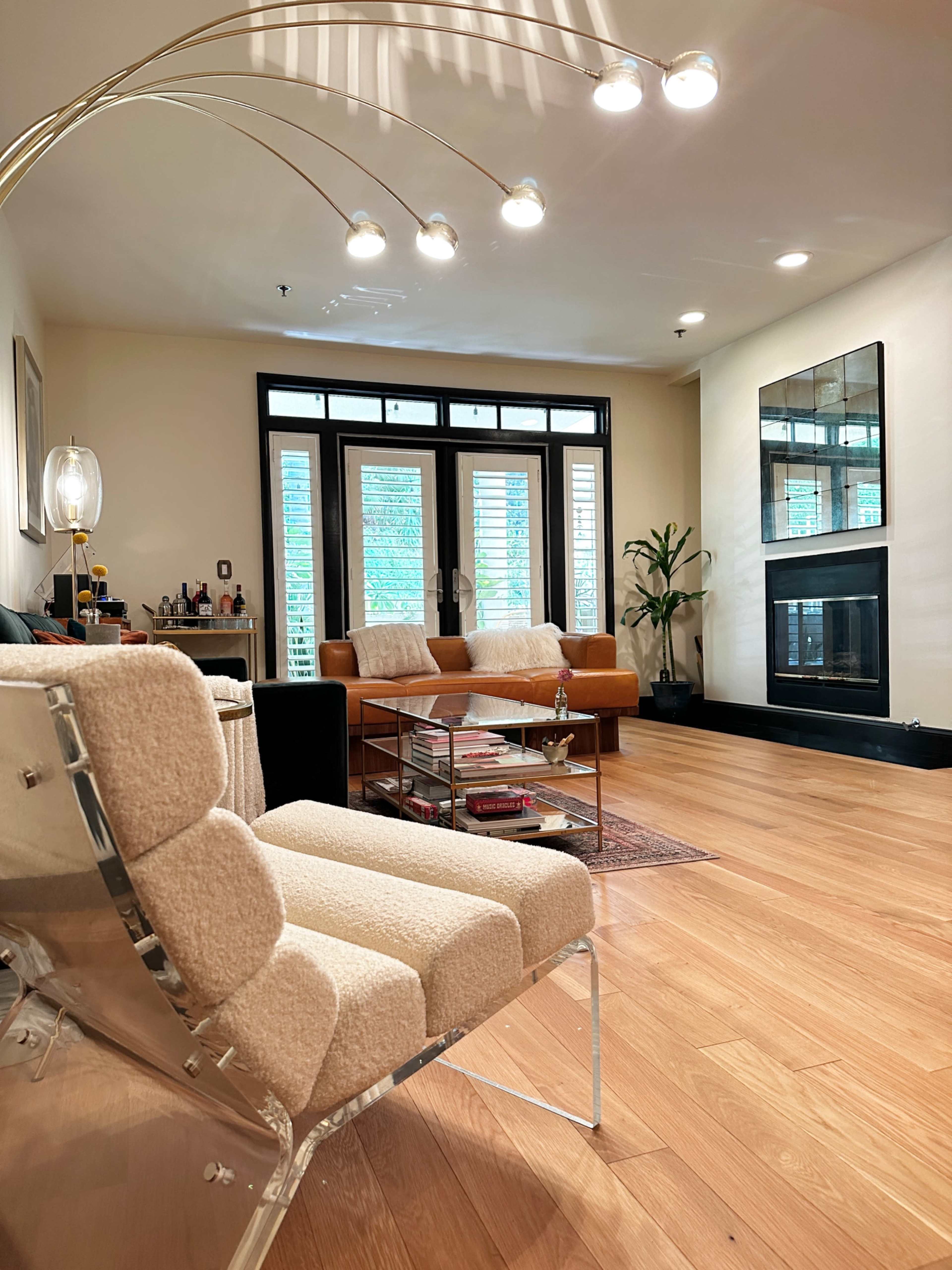 The image shows a cozy living room with a clear view of a modern armchair, a coffee table, a sofa, and a fireplace, with large windows allowing natural light to enter.