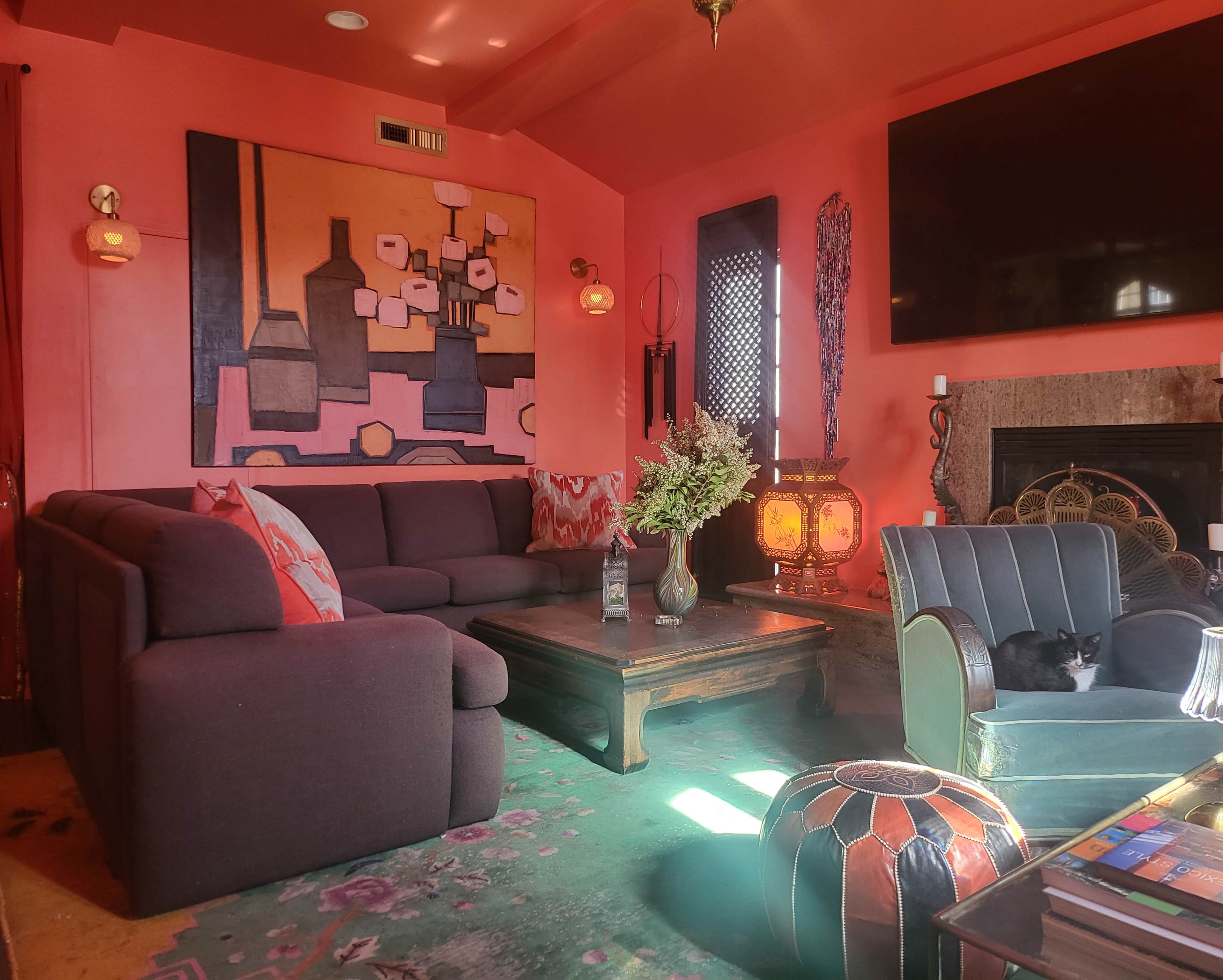 The image shows a cozy living room with a large gray sectional sofa, a wooden coffee table, and decorative elements like a vase of flowers and a black cat resting in an armchair, all set against vibrant coral walls.