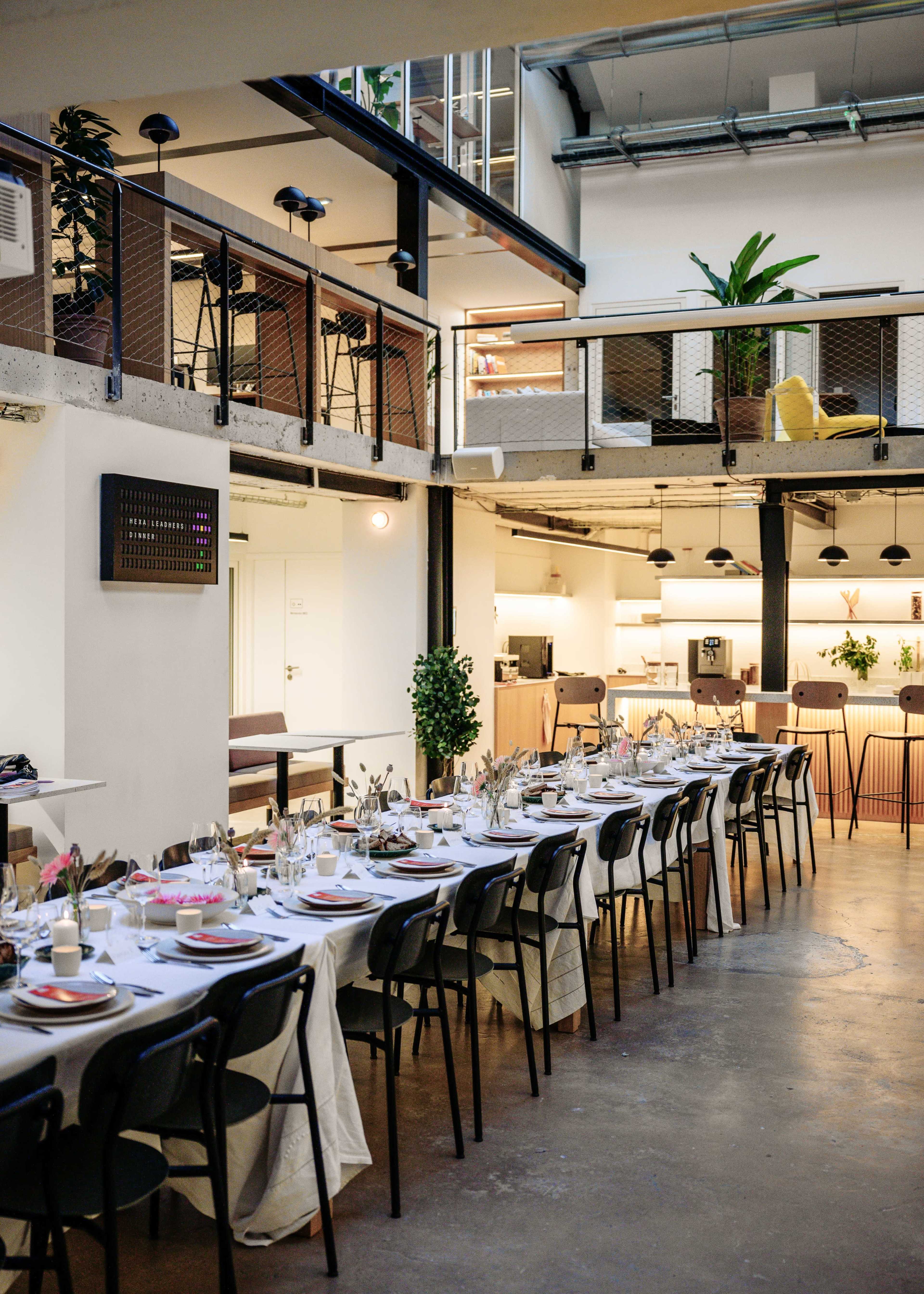 The image shows a dining area with a long table set for a formal meal, surrounded by a modern, open space featuring high ceilings and balcony seating.