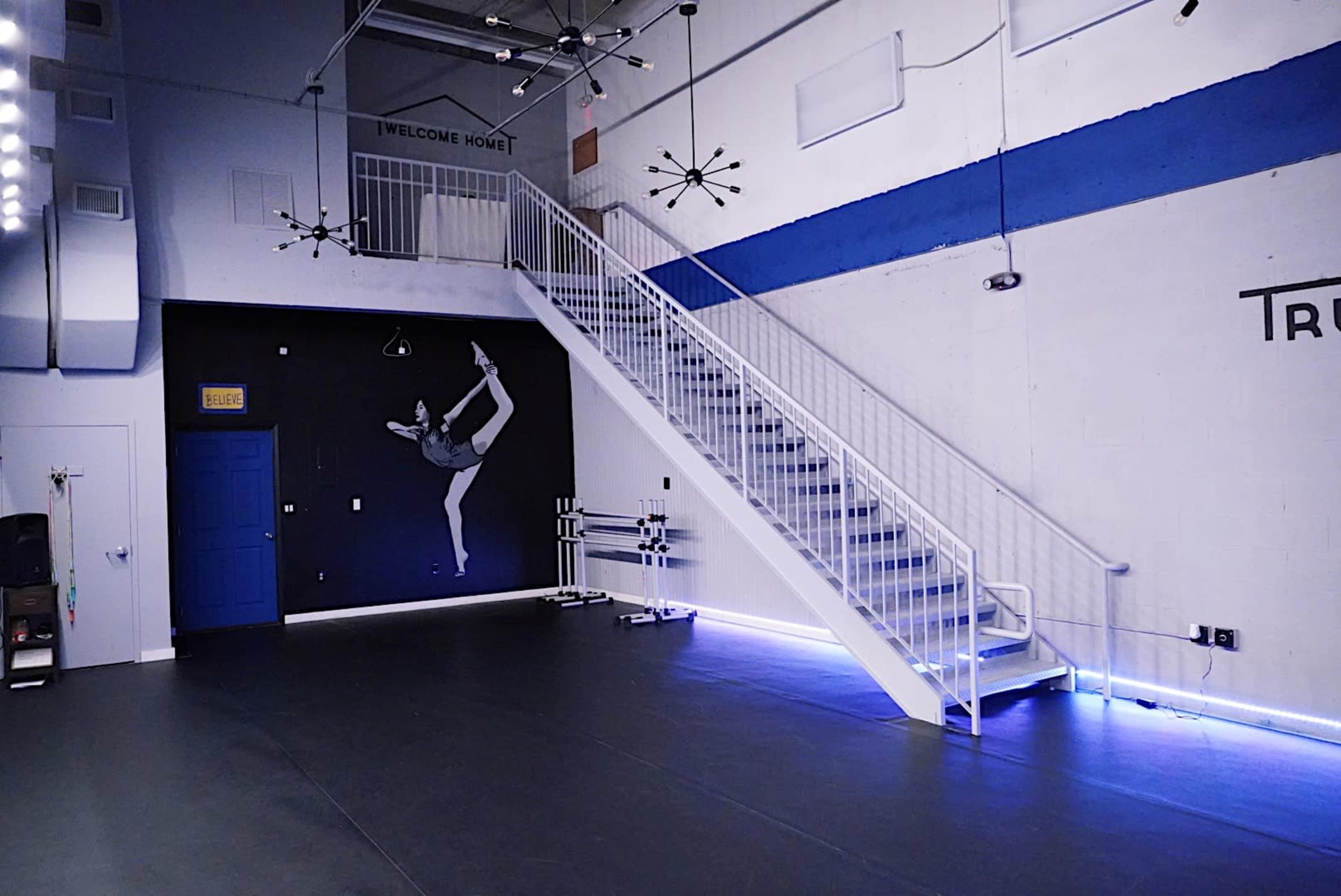 The image shows an interior dance studio featuring a staircase along one wall, a mural of a dancer, and blue and white accents.
