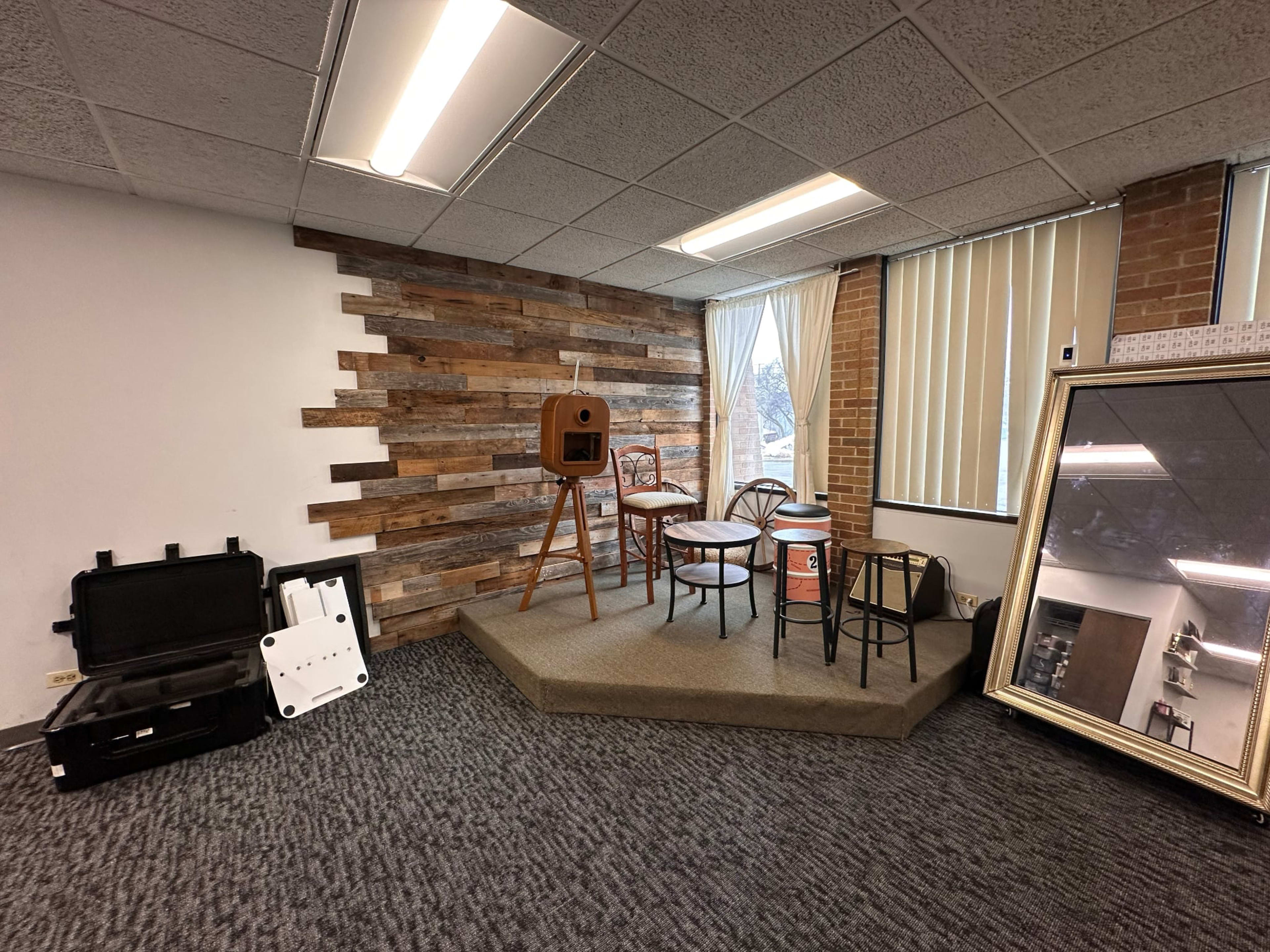The image shows a small stage area with wooden paneling, featuring three chairs and a vintage-style camera, along with a large mirror and a couple of cases on the floor.