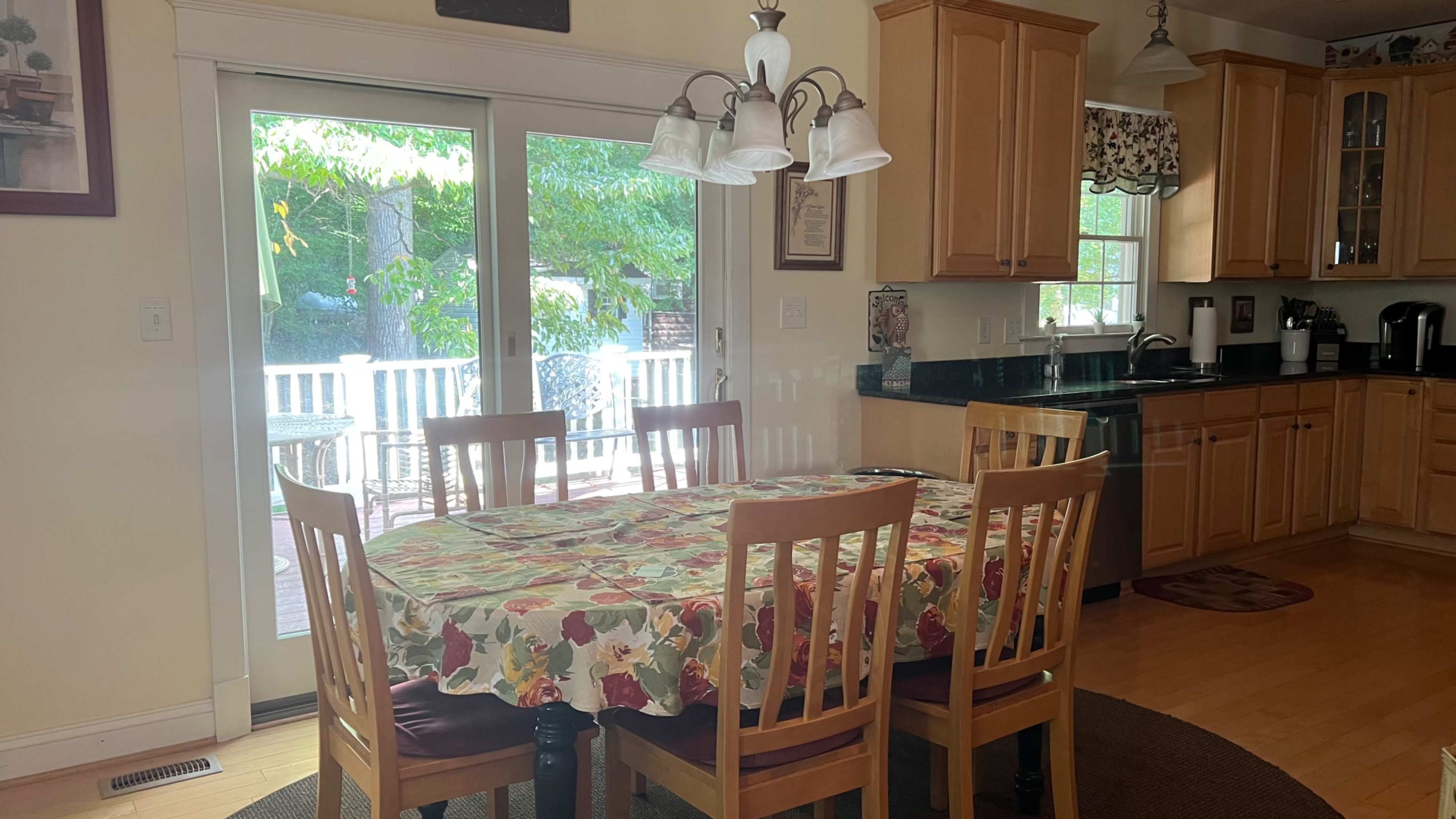 A dining area features a wooden table covered with a floral tablecloth surrounded by six chairs, with sliding glass doors leading to a deck in the background.