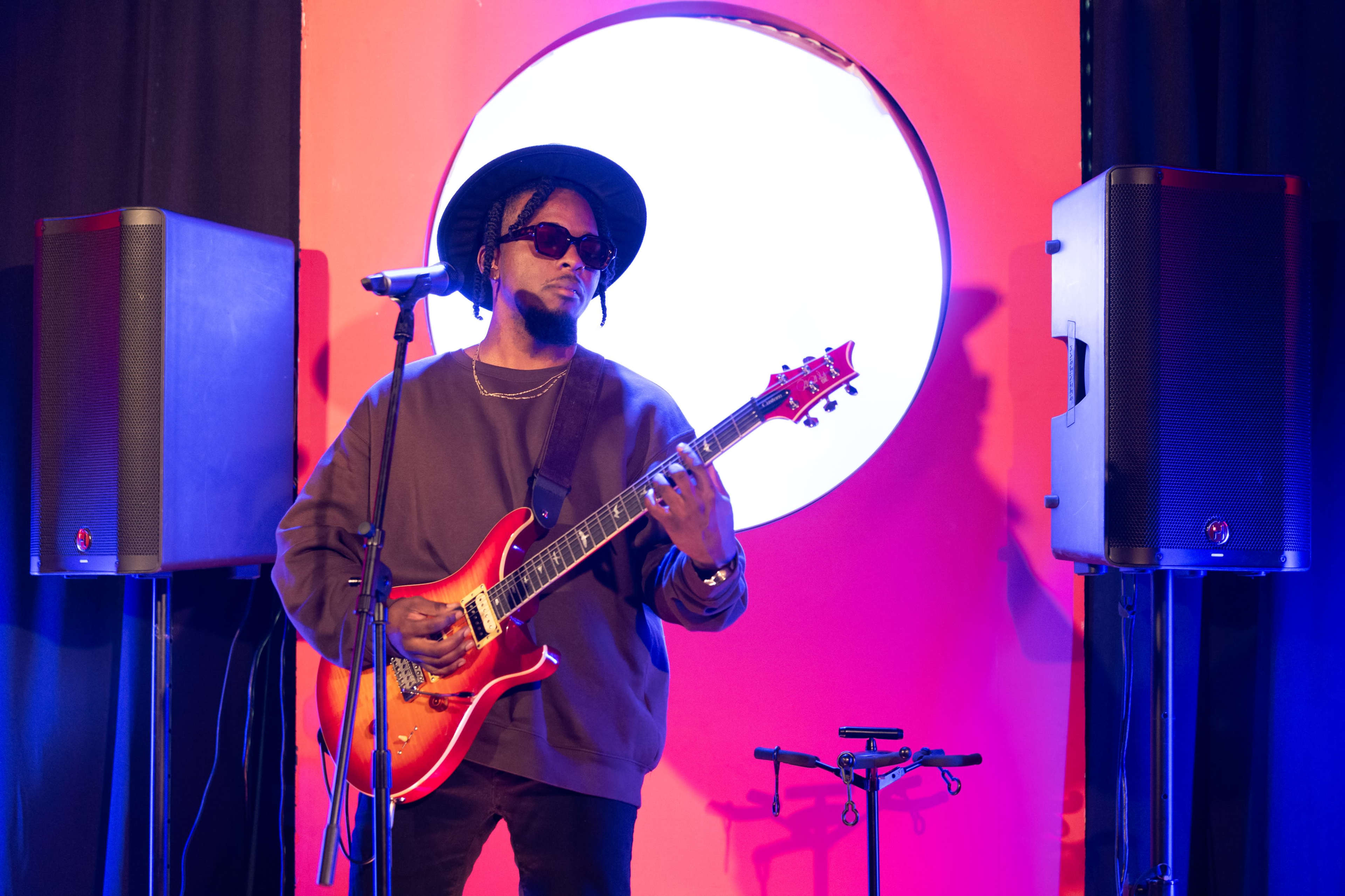 A musician wearing a hat and sunglasses plays an electric guitar on stage, with large speakers and a circular backdrop in the scene.