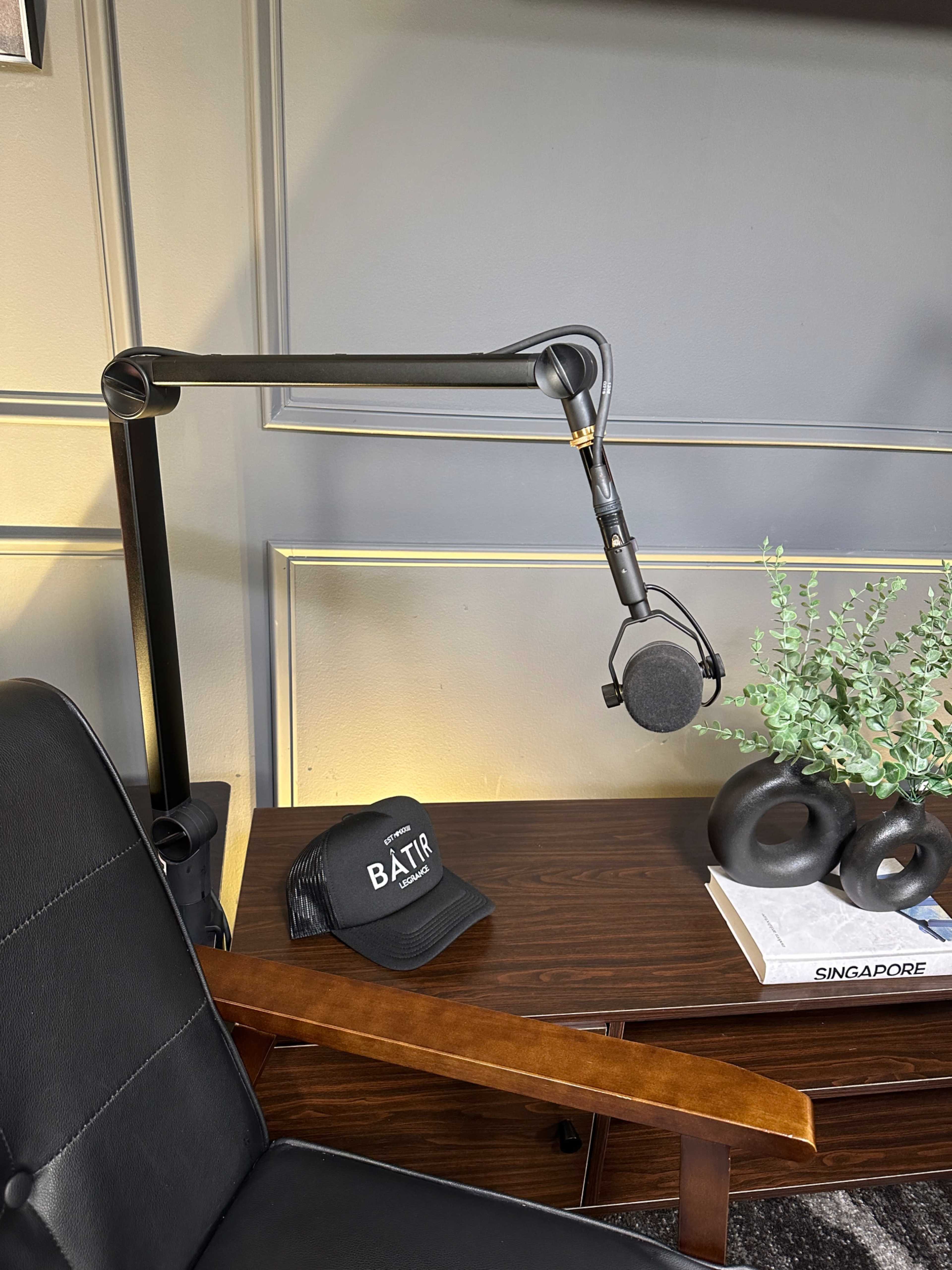 The image shows a black microphone mounted on an adjustable arm next to a dark chair and a wooden table with a hat and decorative items.