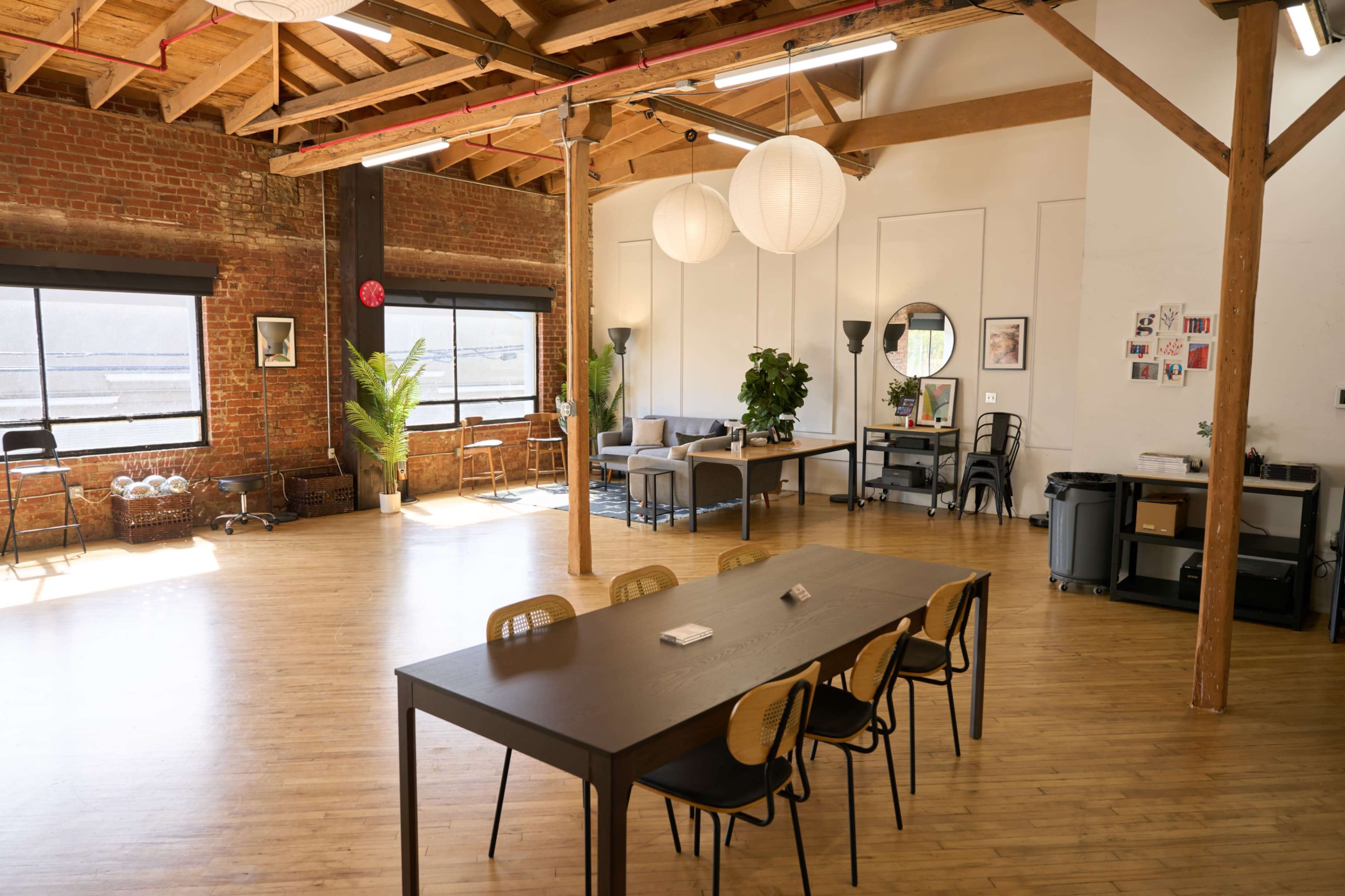 The image shows a spacious, well-lit interior with wooden beams, a long table surrounded by chairs, and various seating areas against a backdrop of large windows.