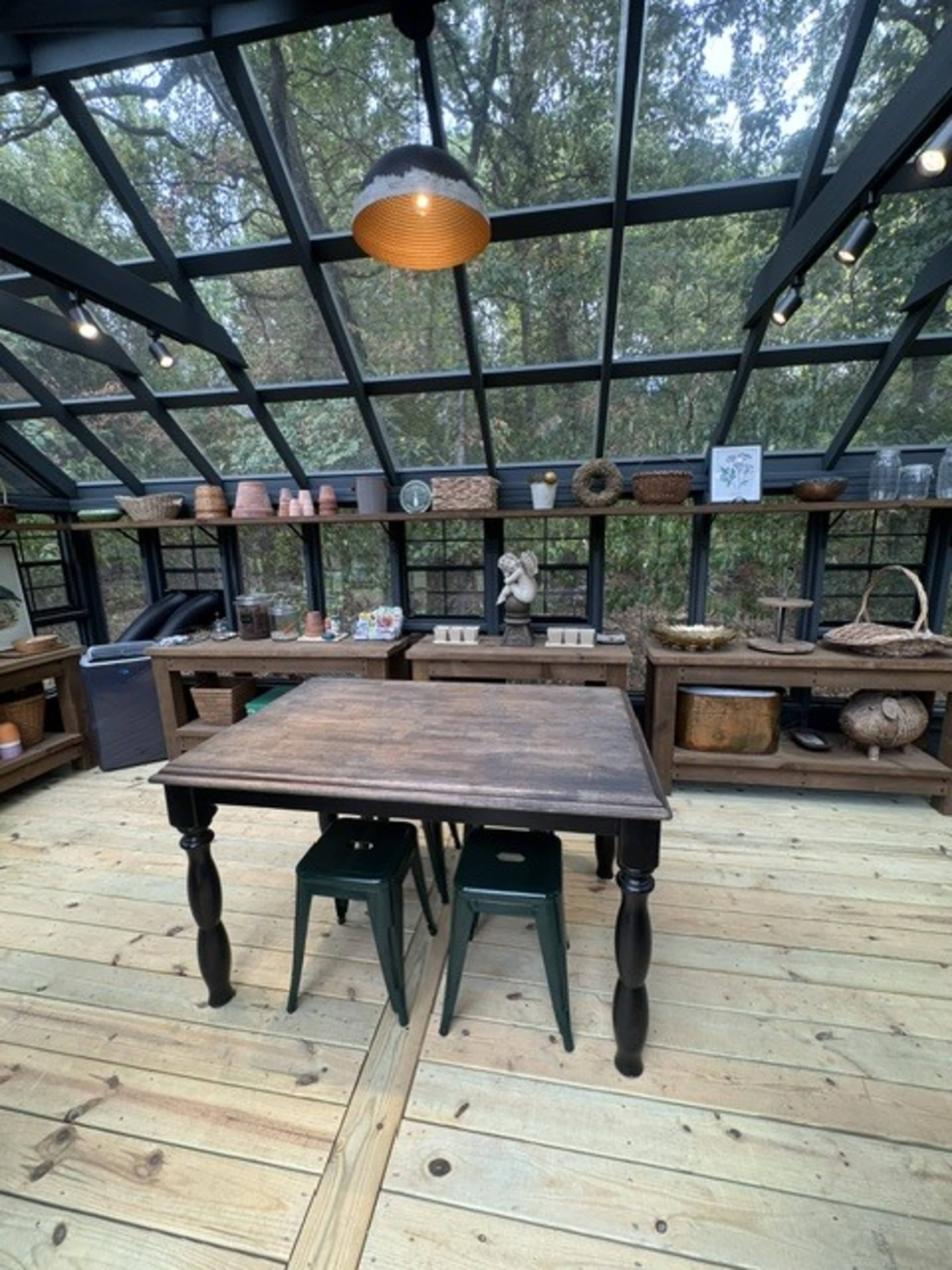 The image shows a wooden table with two green stools situated in a glass-enclosed space filled with various containers and decor items along the walls.