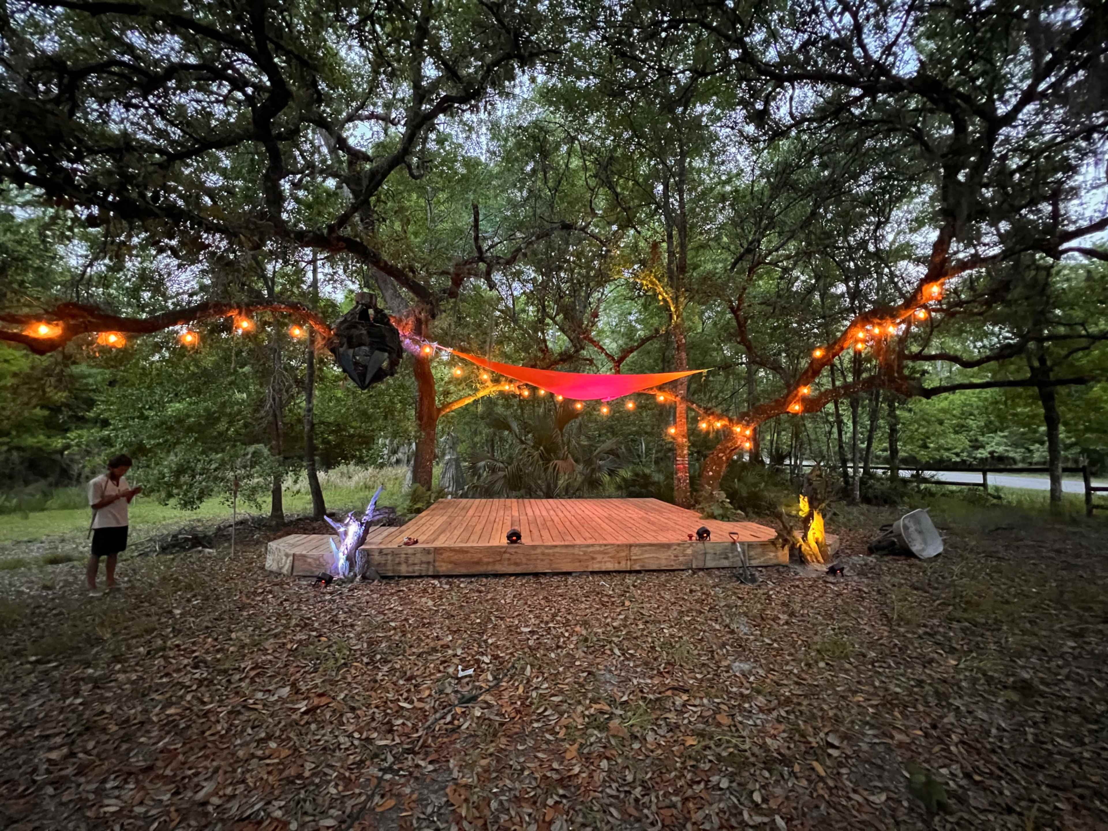 A wooden stage is set under trees adorned with string lights and colorful fabric, surrounded by a natural forest backdrop.