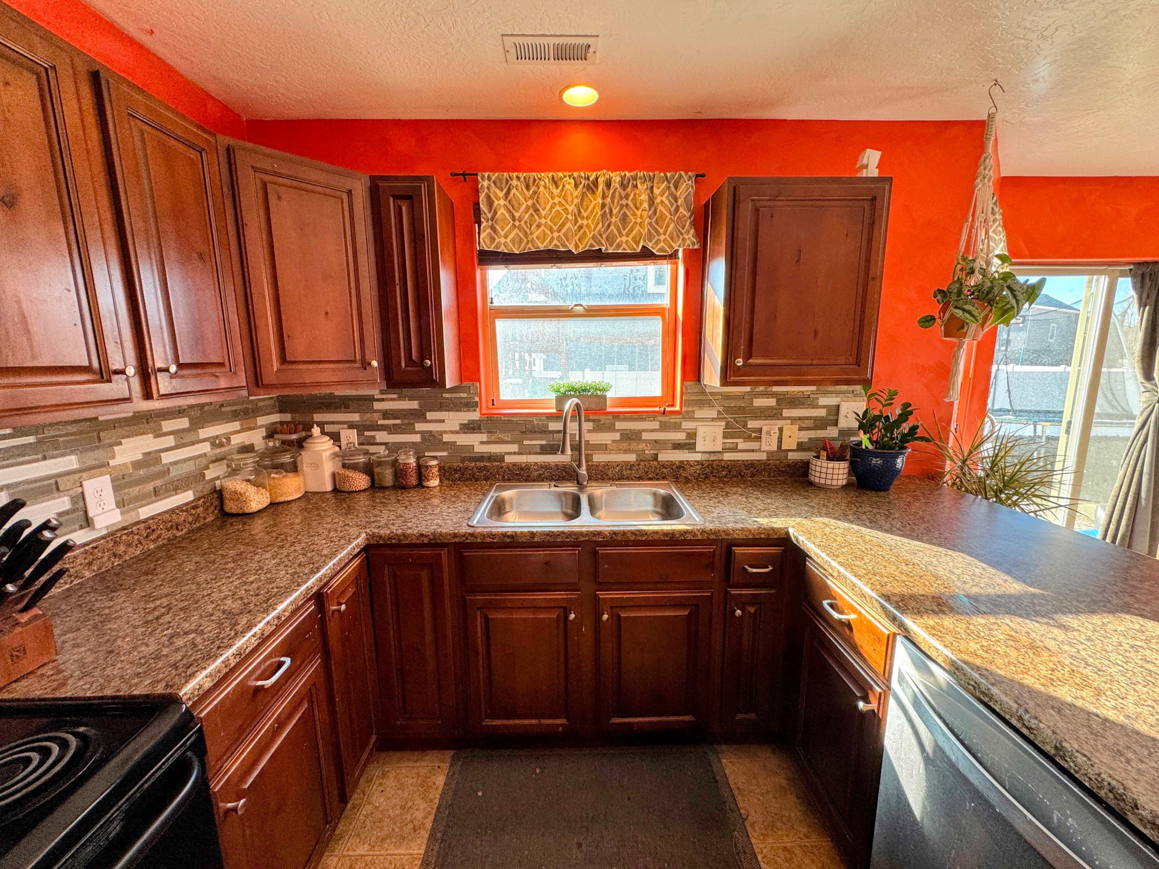 The image shows a kitchen with wooden cabinets, a double sink, a granite countertop, and a vibrant red wall with a window above the sink.